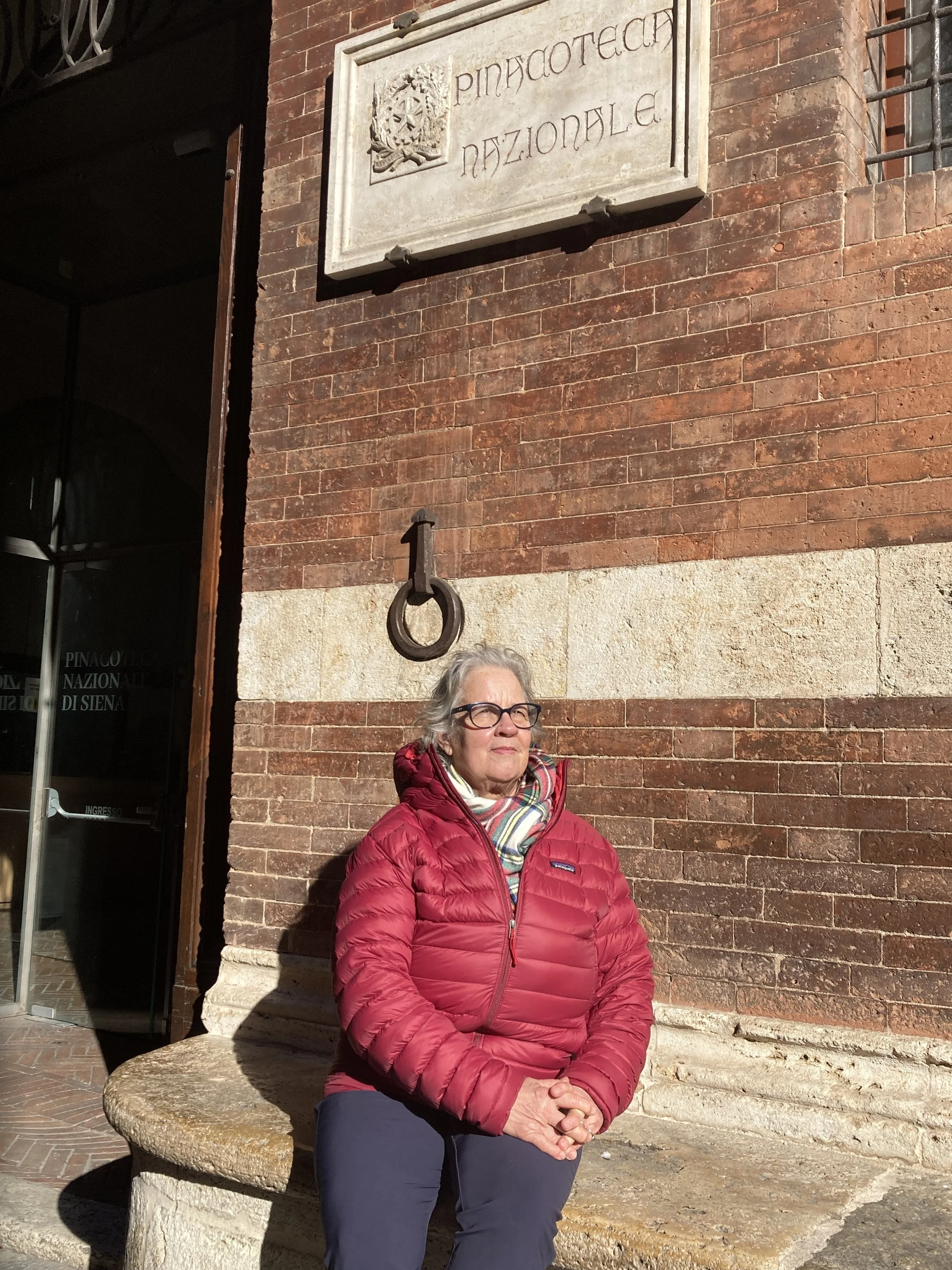 An elderly woman in a red jacket and glasses sitting on a stone bench outside a building with brick walls and a large metal hook on the wall.