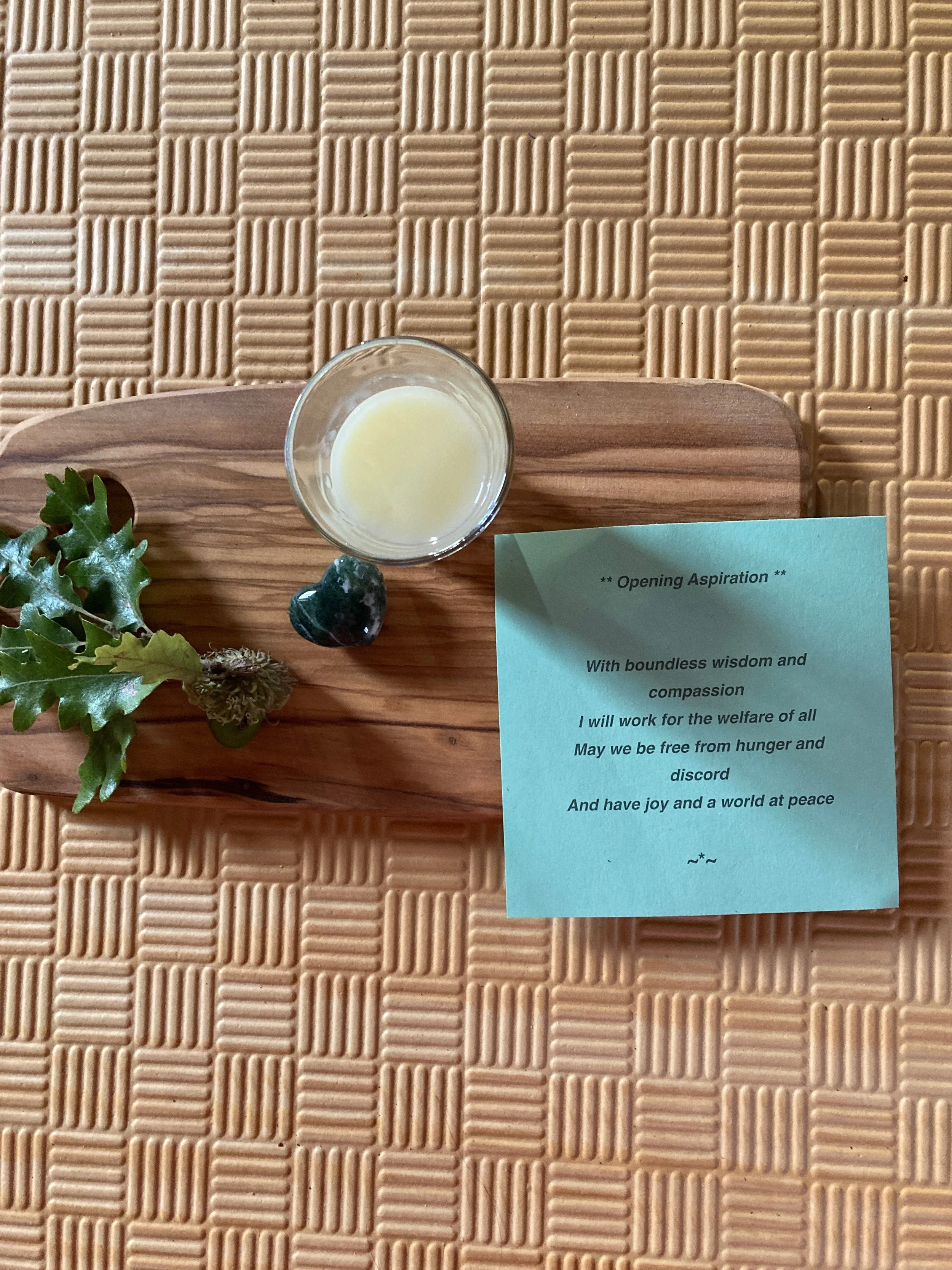 Small glass of a light-colored liquid, possibly tea or honey, on a wooden tray with a sprig of green leaves and a black stone. A blue note with a poem or aspiration is next to the tray, placed on a textured beige surface.