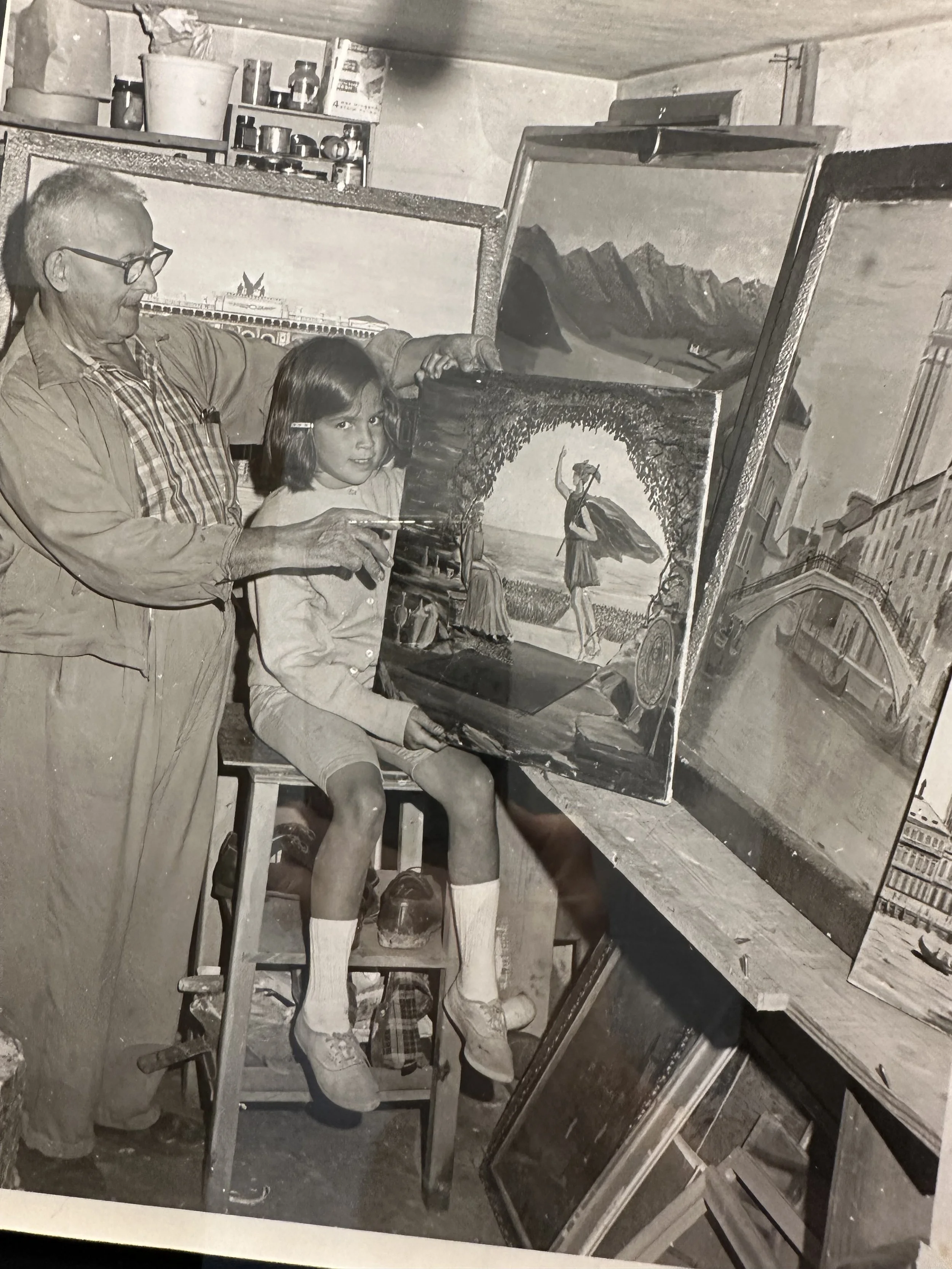 An older man and a young girl are in an art studio, working on a painting together. The girl is seated on a stool, looking at the camera, while the man points at the painting with a brush. There are several paintings and art supplies around them.