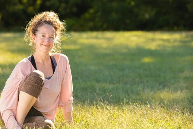 A woman sitting on grass in a park during daytime, smiling at the camera.