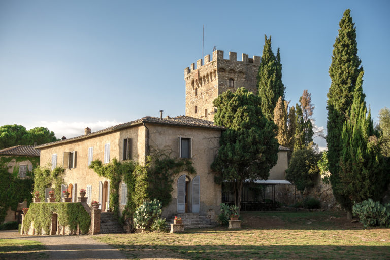 A stone castle tower with battlements surrounded by tall cypress and other trees, with a rustic villa in front, under a clear blue sky.