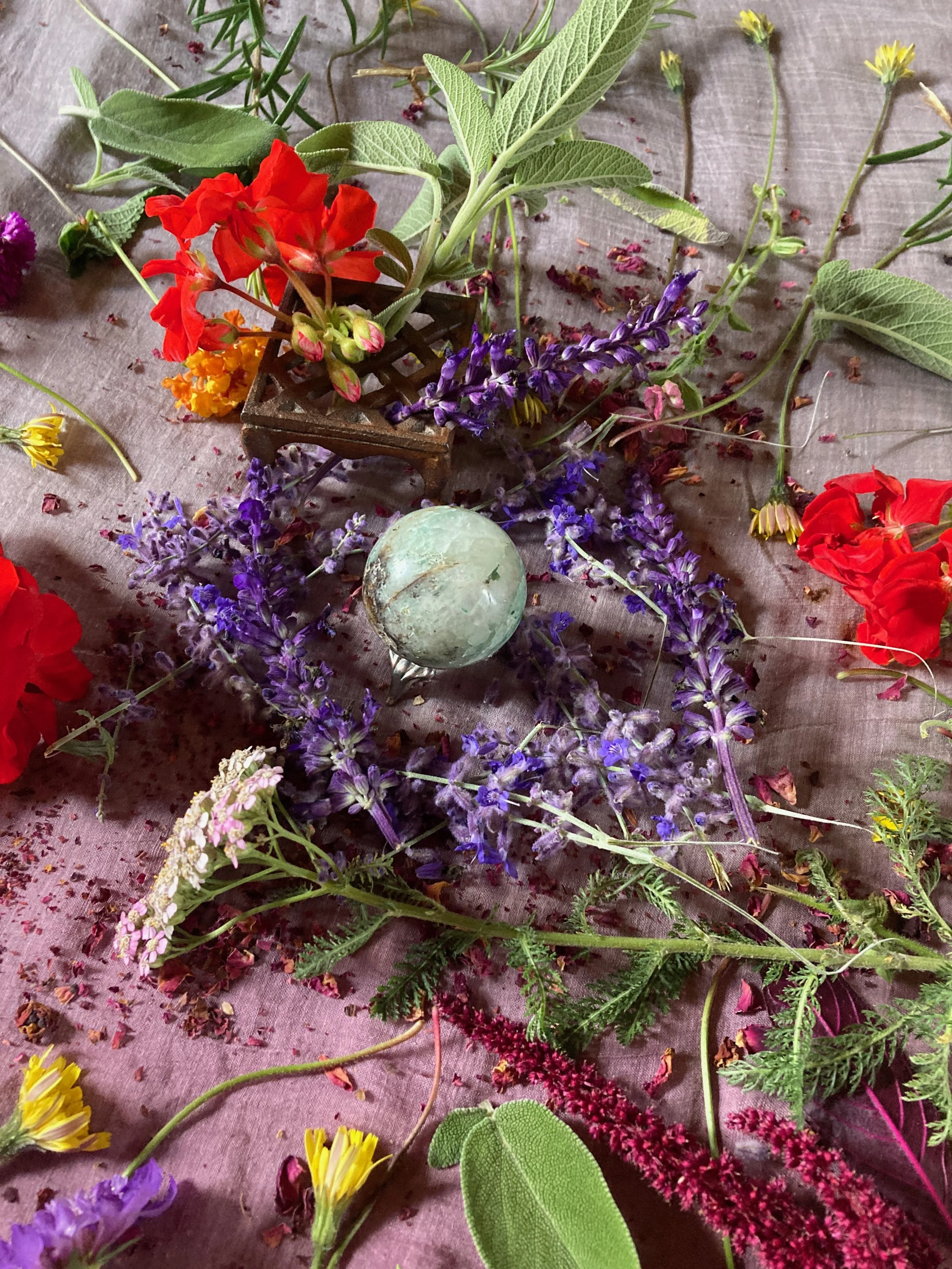 Colorful assortment of fresh flowers and greenery laid on a purple fabric surface, including purple lavender, red, yellow, and pink blooms with various green leaves, and a round green and white marble.