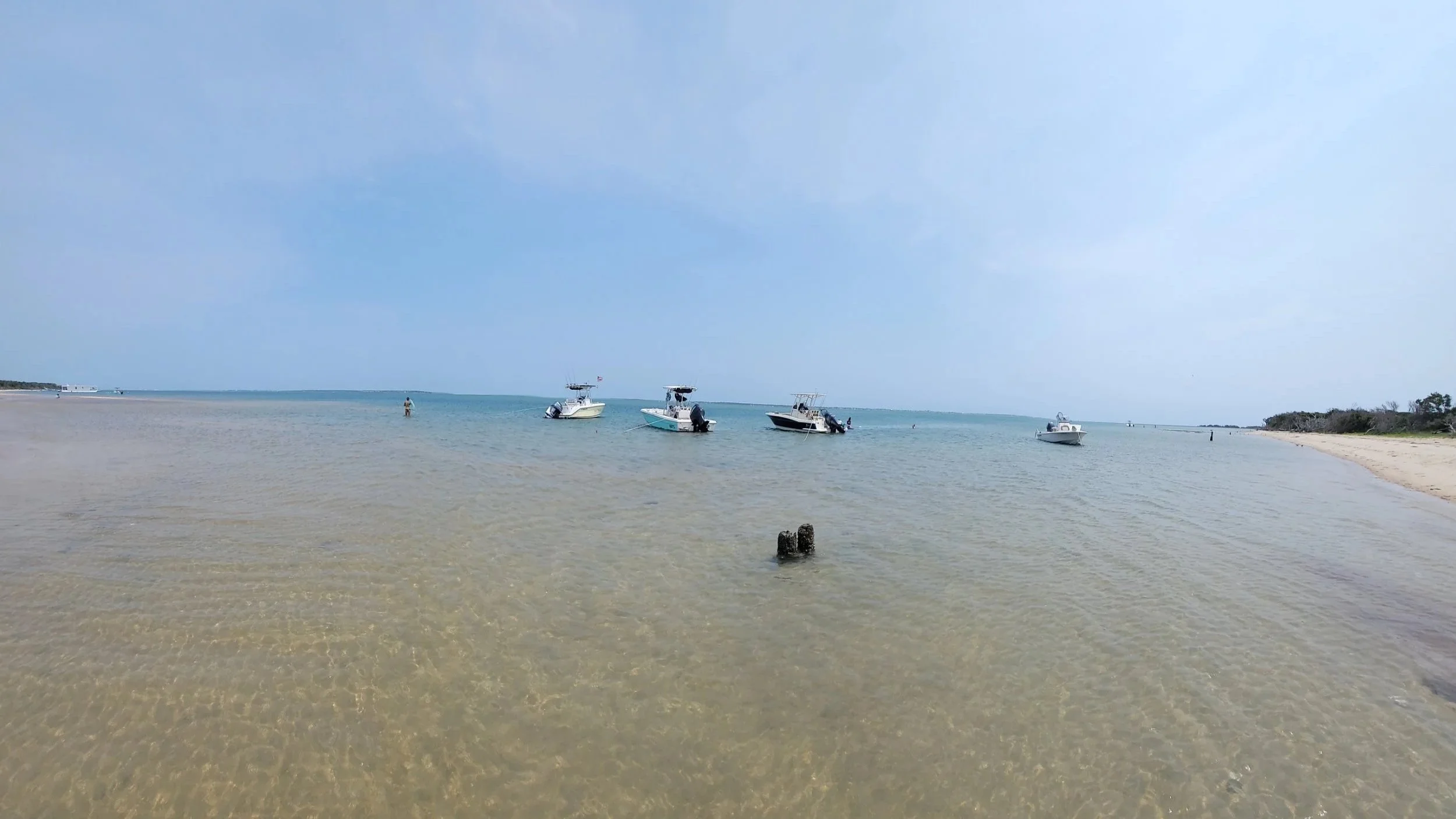 Calm ocean water with five boats anchored near the shore, a sandy beach on the right, and a clear sky.