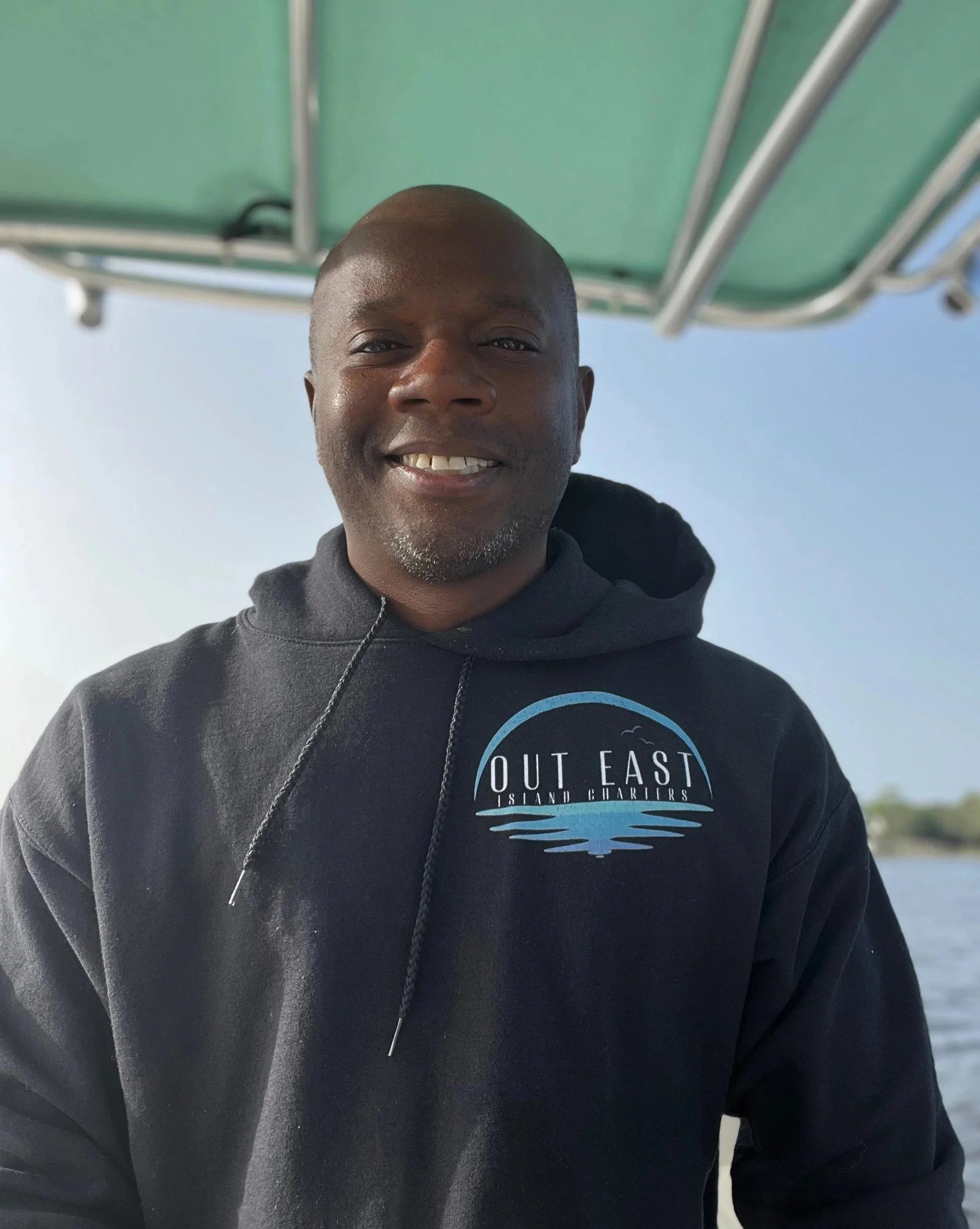 A man smiling on a boat, wearing a black hoodie with a logo that says Out East Island Charters, with a body of water and trees in the background.