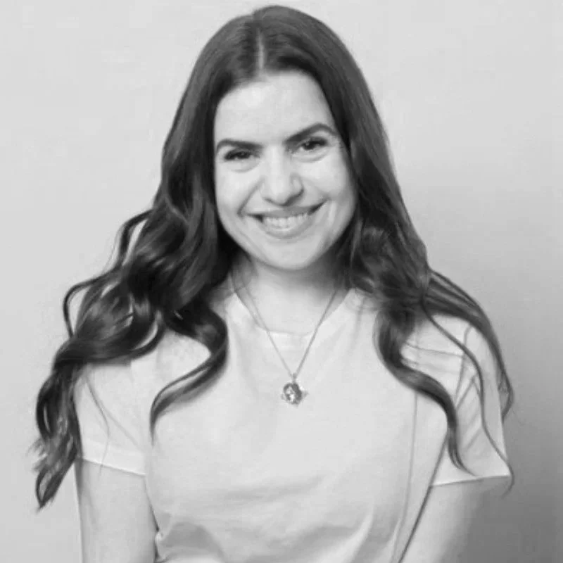 Black and white photo of Julia Falci smiling. She has long wavy hair, wearing a light-colored top and a necklace, against a plain background.
