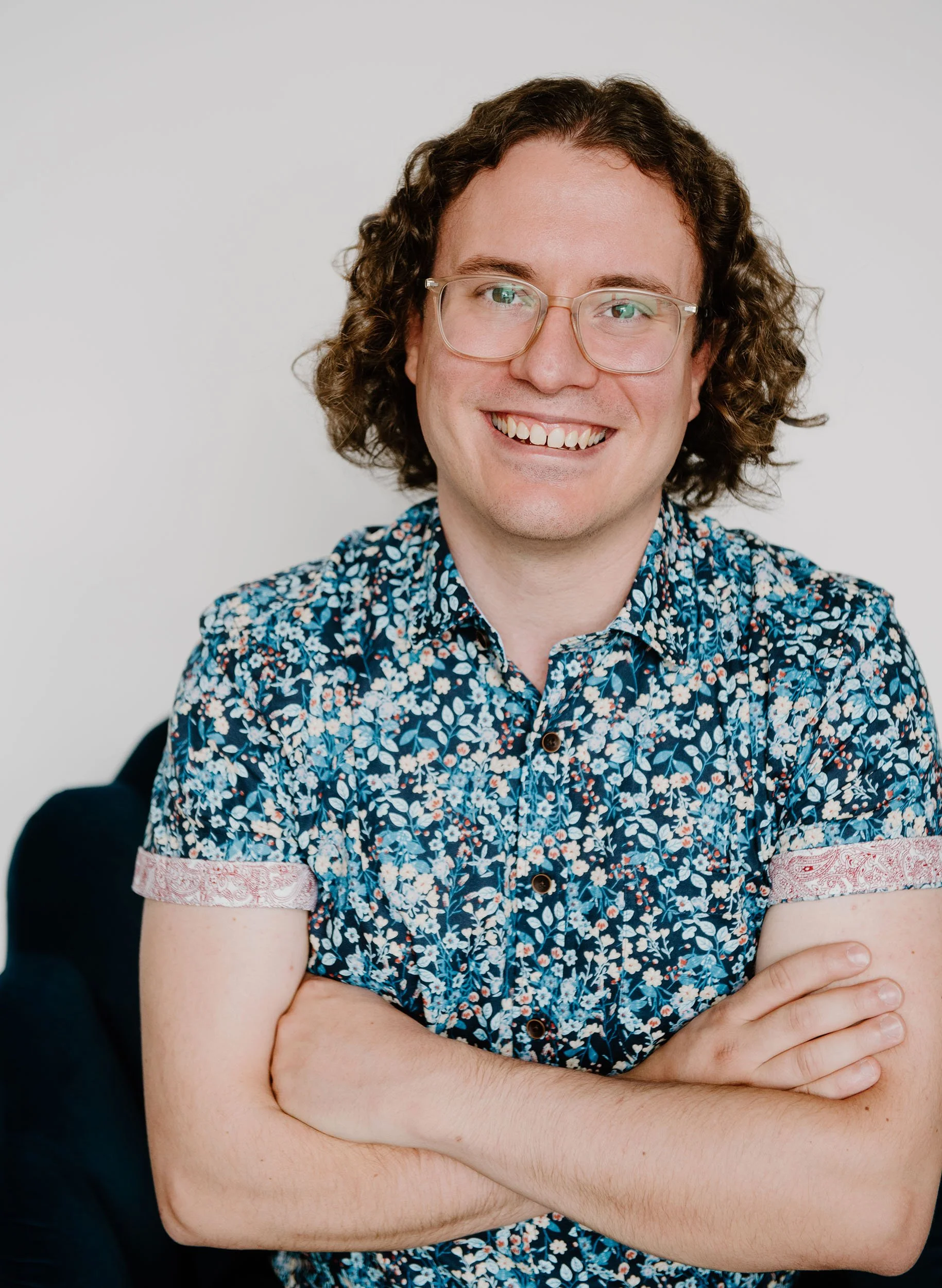 Anthony Cenerini is a young man with curly brown hair, glasses, and a big smile, wearing a short-sleeved, blue floral shirt, standing against a white background.