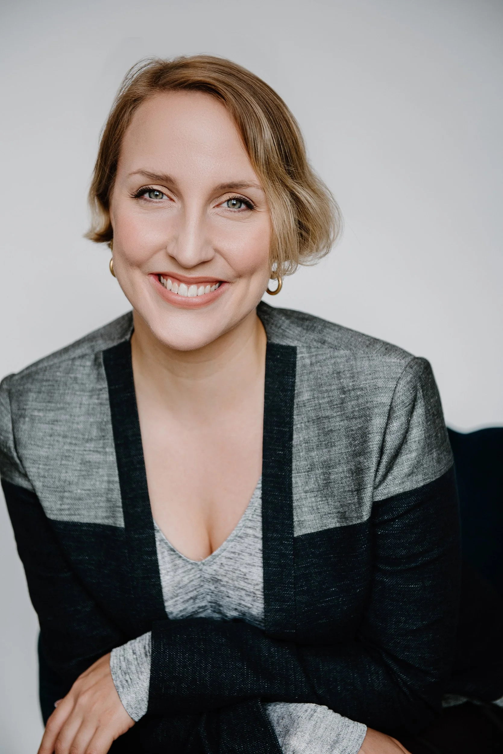 Victoria Cowan is a smiling woman with short blonde hair, wearing a gray and black blazer and a light gray top, sitting against a plain background.