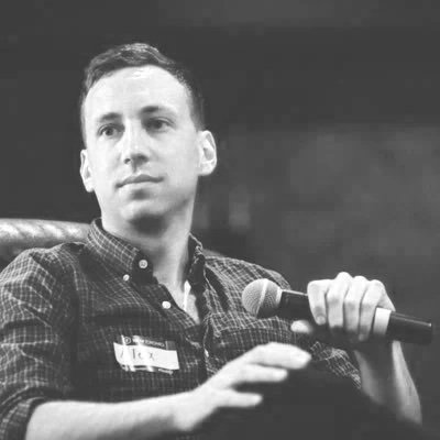 Alex Blumenstein, young man with short hair and a checkered shirt is holding a microphone, sitting in front of a dark background.