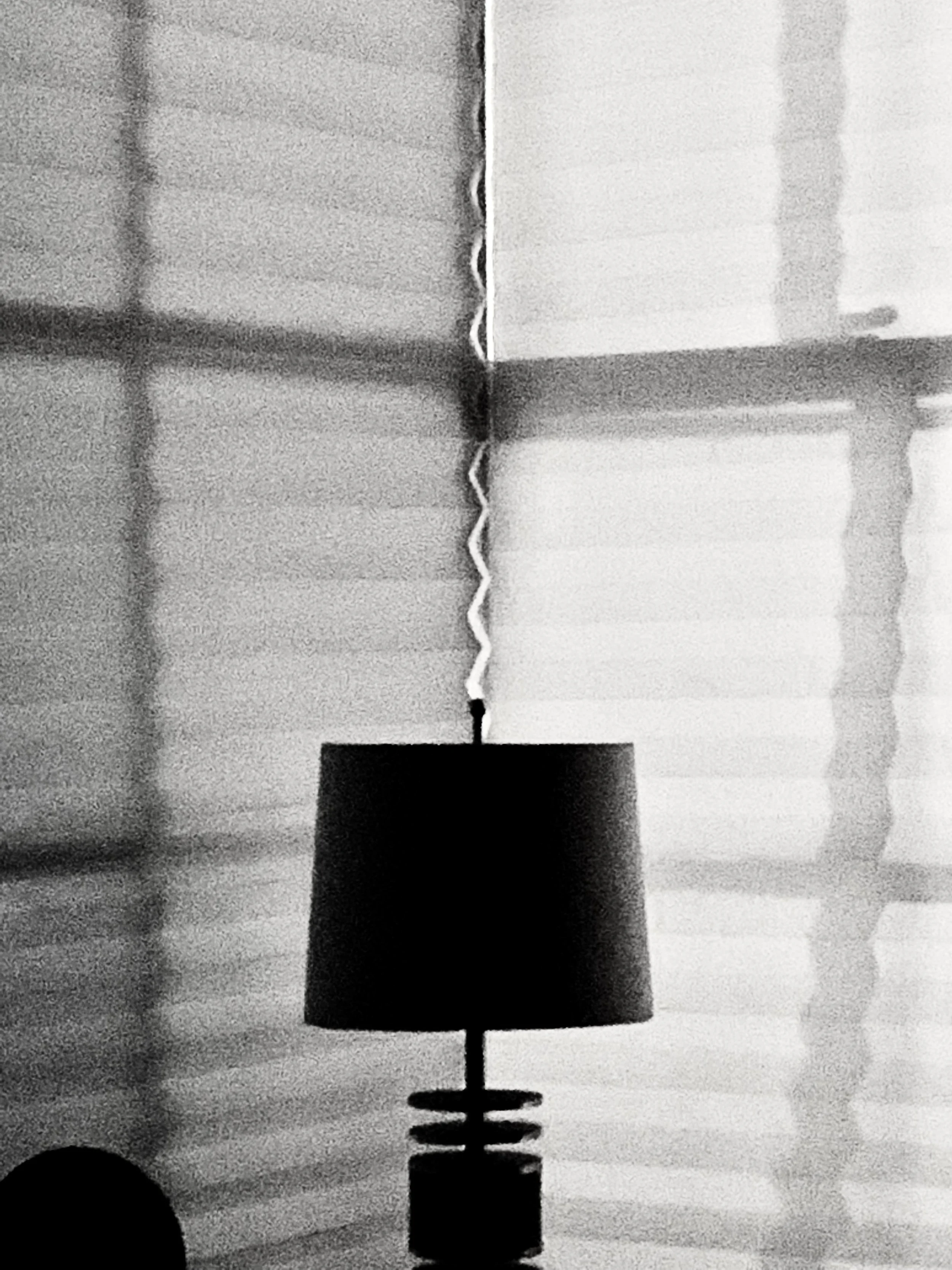 Black lampshade on a table with shadow patterns cast on window blinds in the background.