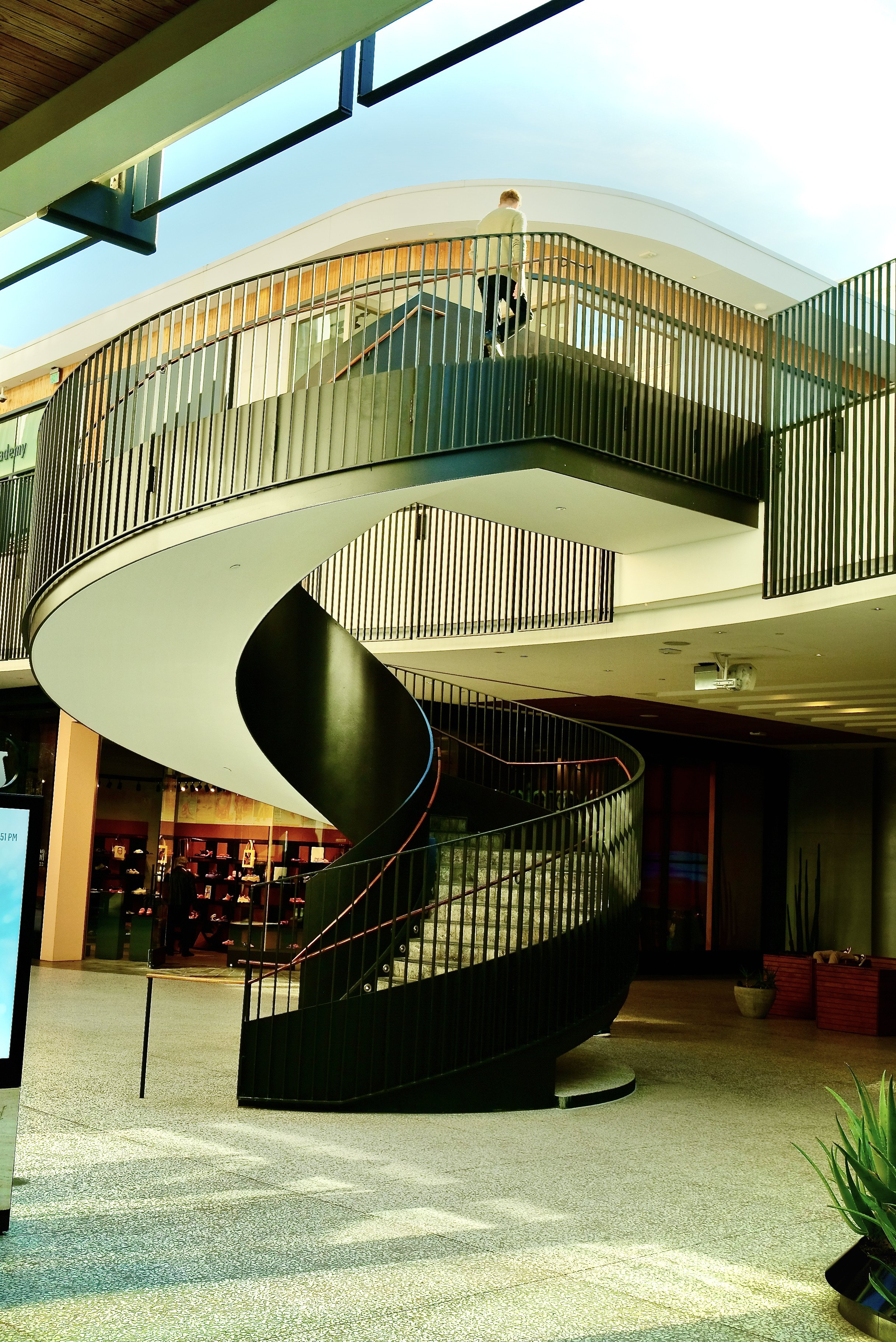 An indoor public space featuring a black spiral staircase with a metal railing, leading to an upper level with a person walking on the balcony under a bright blue sky visible through the ceiling.