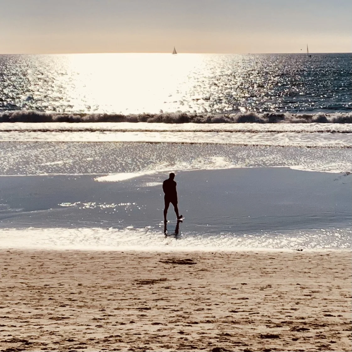 Person standing on the wet sand at the beach, looking at the ocean during sunset or sunrise, with sailboats visible on the horizon and gentle waves.