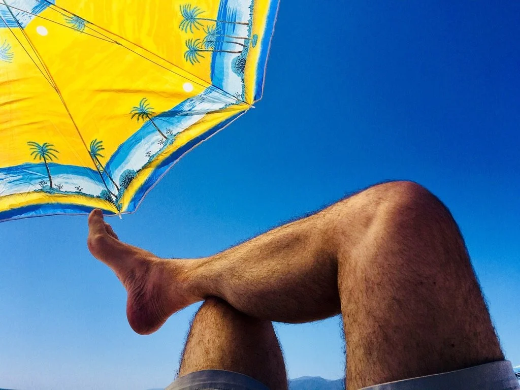 A person's legs with one knee bent, lying on their back, holding a yellow and blue beach umbrella against a clear blue sky.