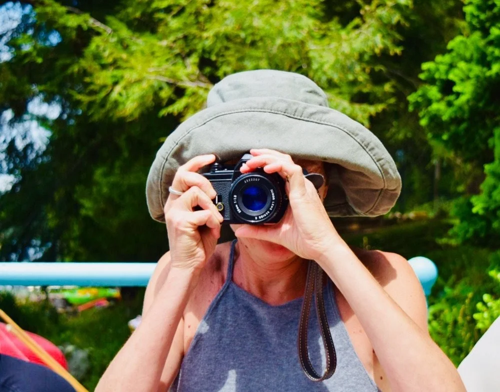Person wearing a large sun hat and a tank top, holding a camera up to her face, outdoors with green trees in the background.