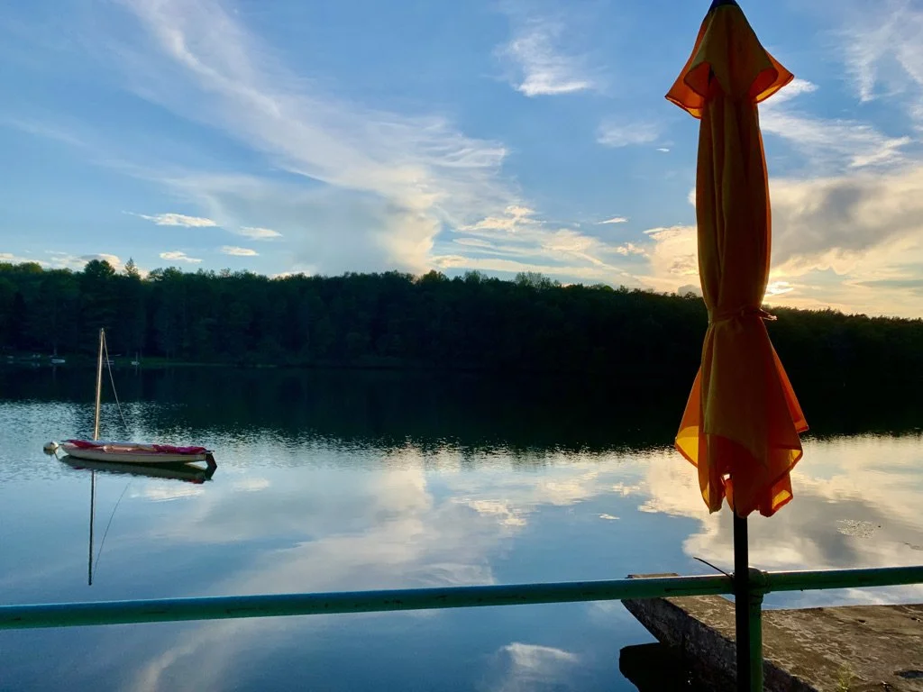 Calm lake with sailboat and a closed orange umbrella on a dock at sunset