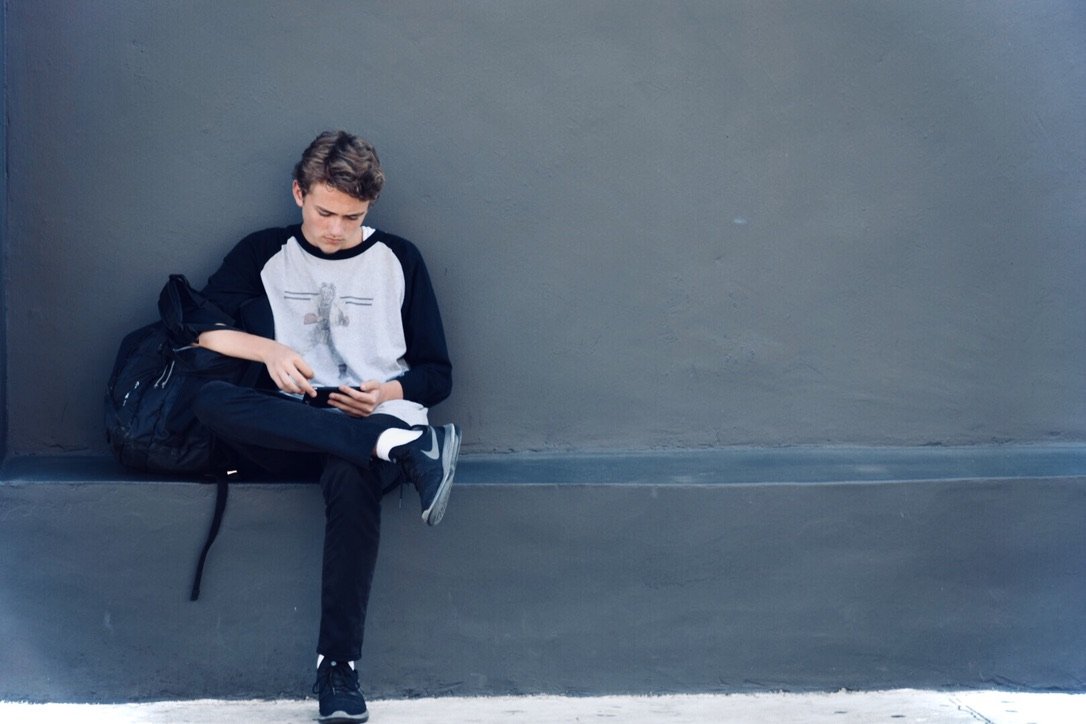 A young boy sitting alone on a bench, looking at his phone, with a backpack beside him, against a plain gray wall.
