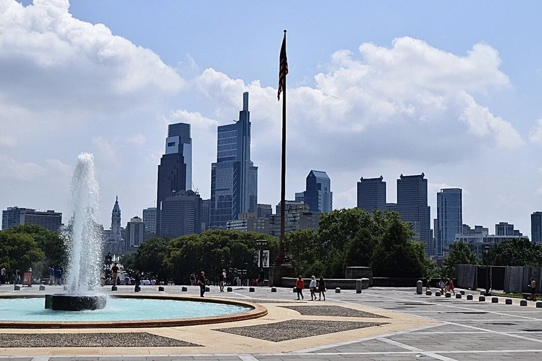 View of Philadelphia skyline with a fountain and American flag in front, on a partly cloudy day.