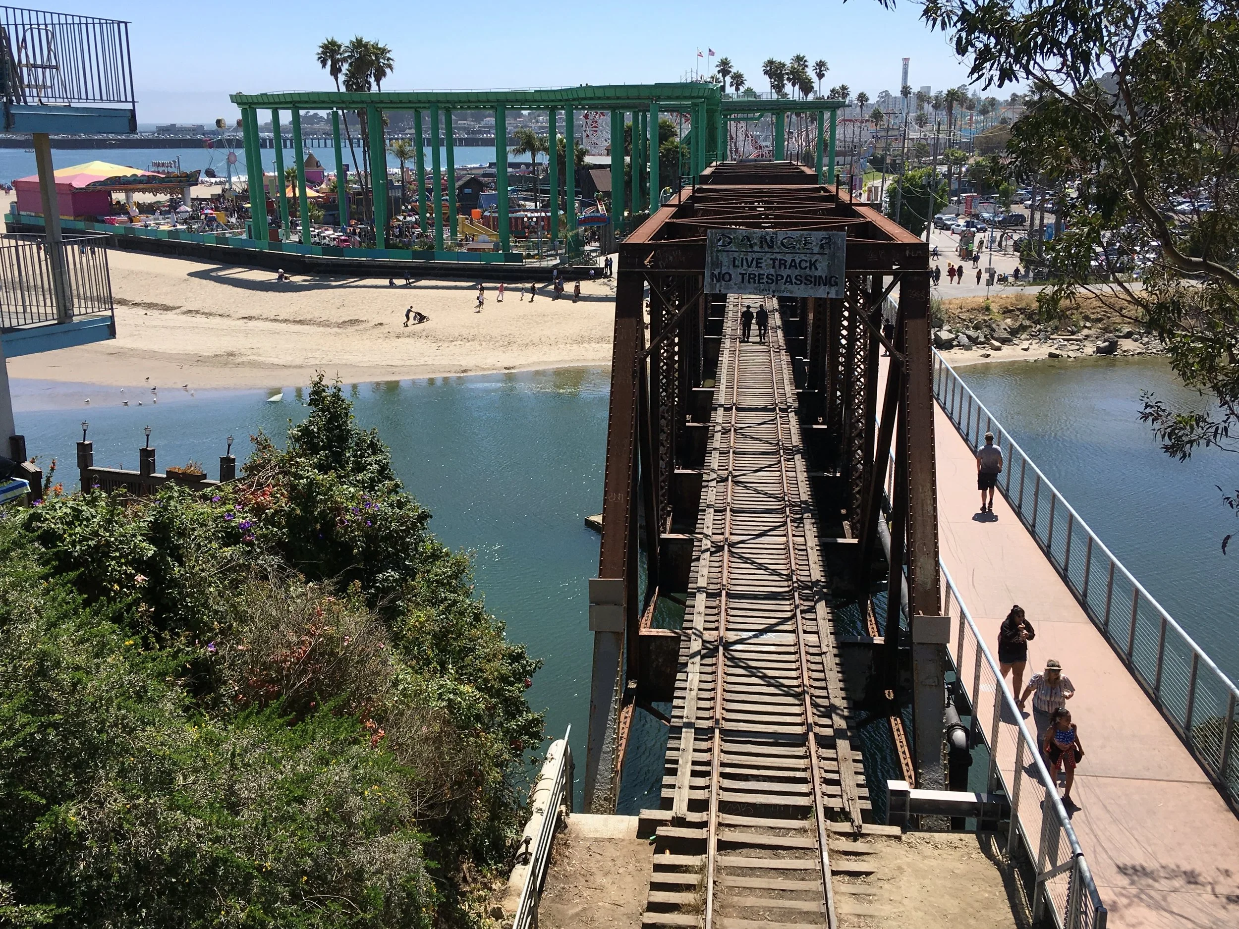 View of a historic rusty railway bridge with a sign that says 'DANGER LIVE TRACK NO TRESPASSING,' over a body of water. On the right, there's a walkway with a few pedestrians. In the background, a sandy beach and an amusement park with rides and colorful structures are visible, along with palm trees and a cityscape in the distance.