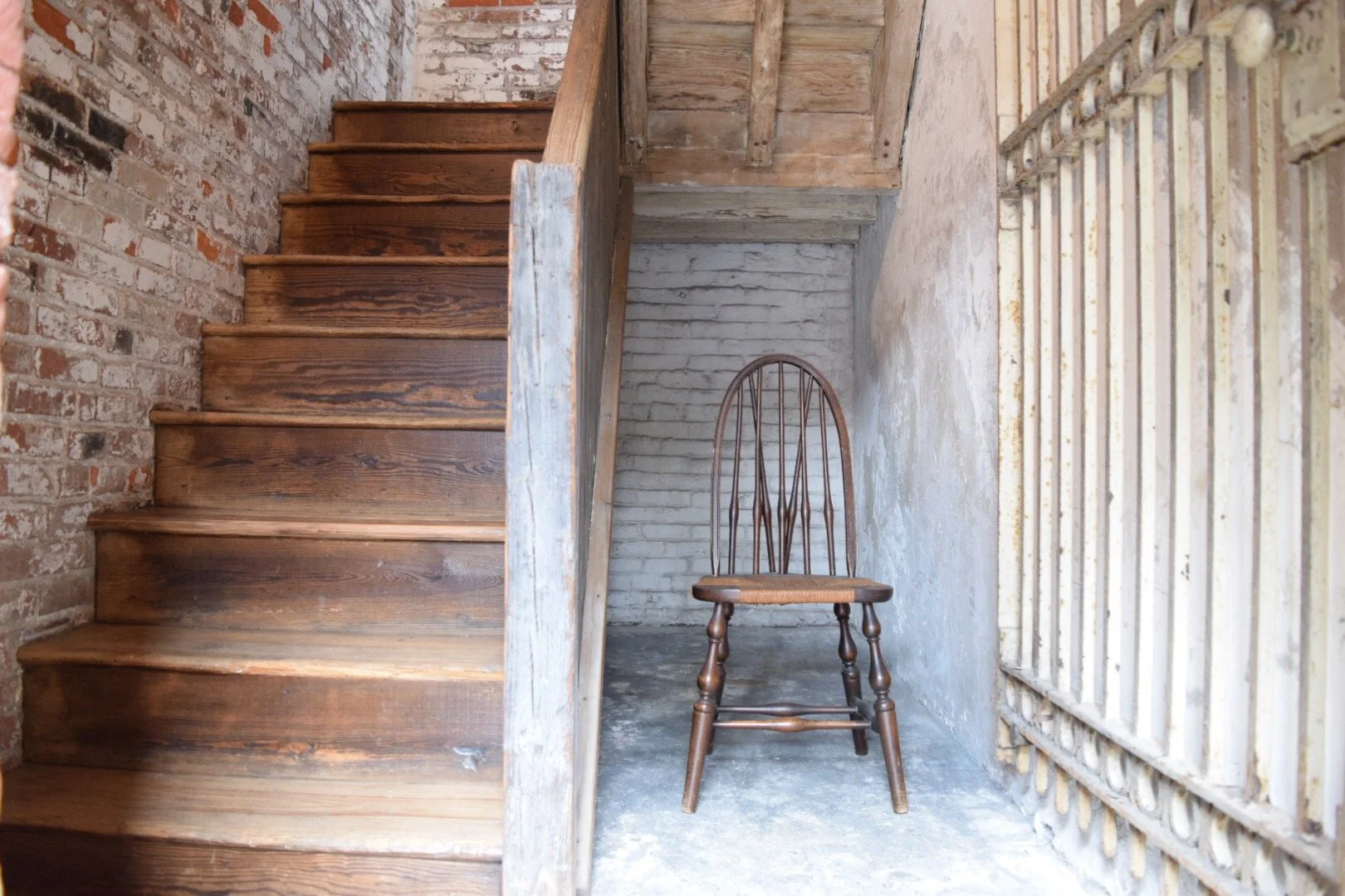A wooden chair with a curved backrest is placed in a small space beneath a staircase, surrounded by brick and concrete walls with a barred window to the right.
