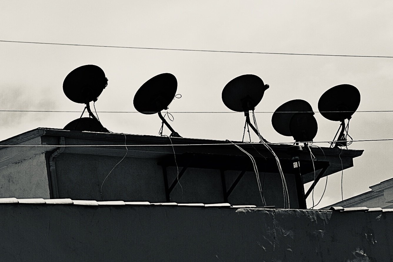 Silhouettes of multiple satellite dishes mounted on the rooftop of a building against a cloudy sky.