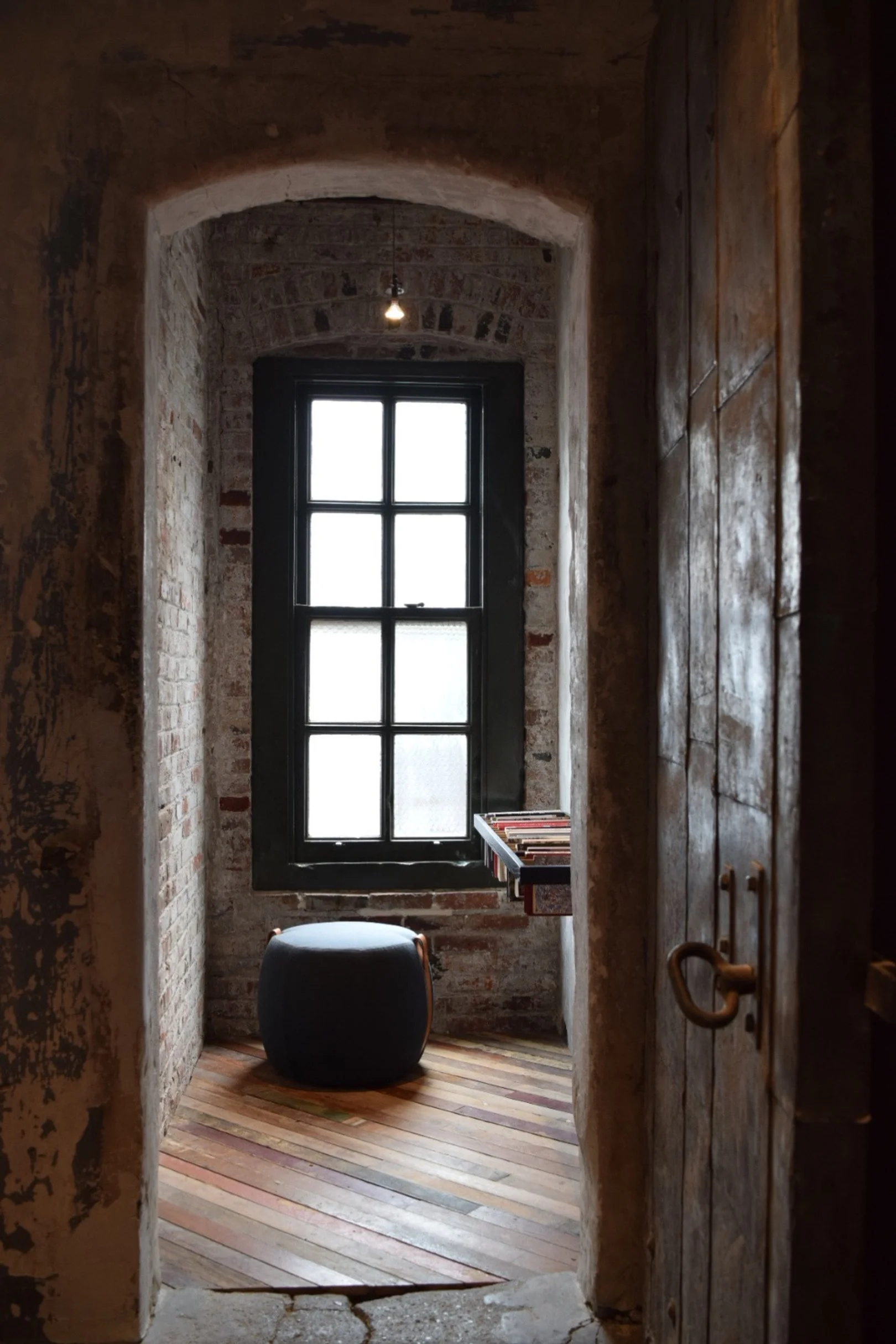 A small room with an arched brick window, a black window frame, a small black ottoman, and a wooden floor, seen through a doorway with a wooden door.