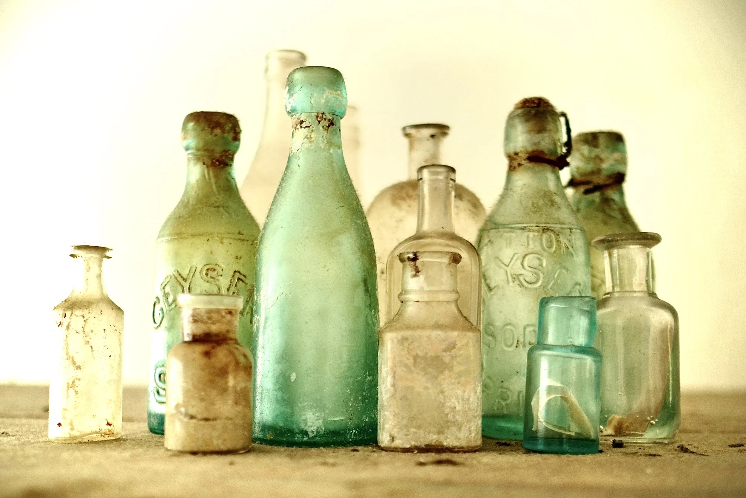 A collection of old glass bottles in various sizes and shades of green, clear, and amber, arranged on a wooden surface with a plain beige background.