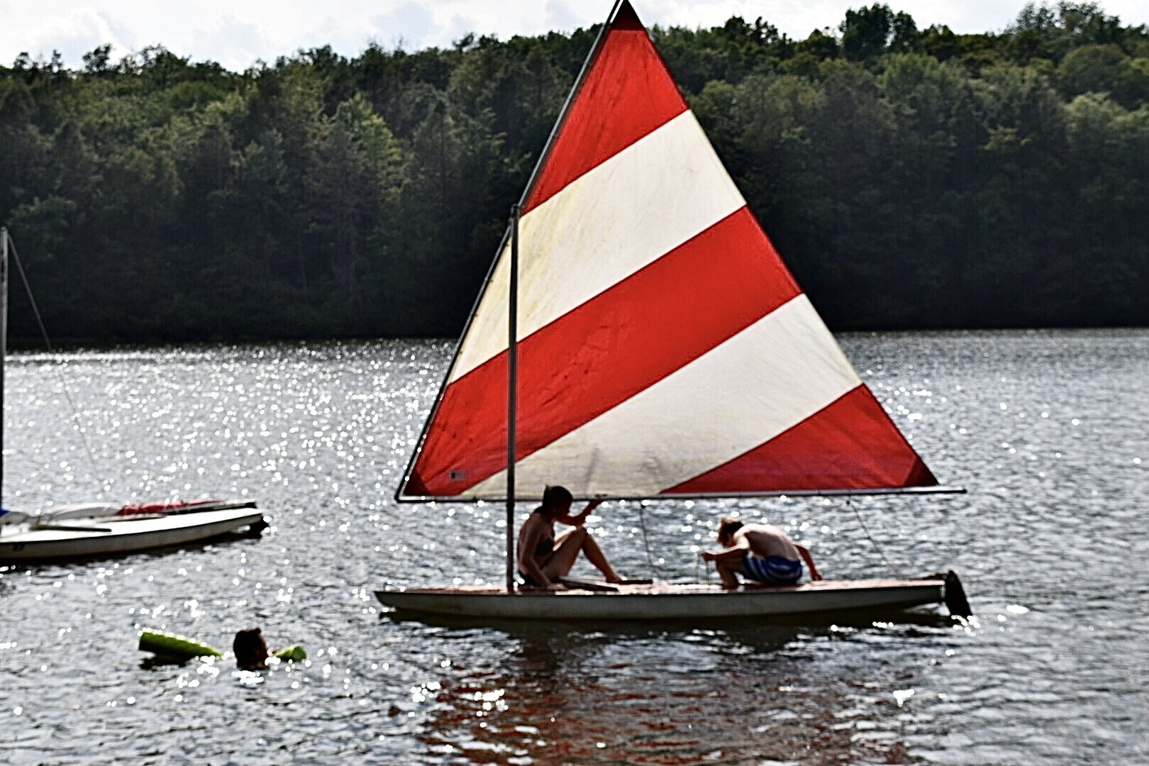 Two children on a small sailboat with a red and white striped sail, floating on a lake with a wooded shoreline in the background