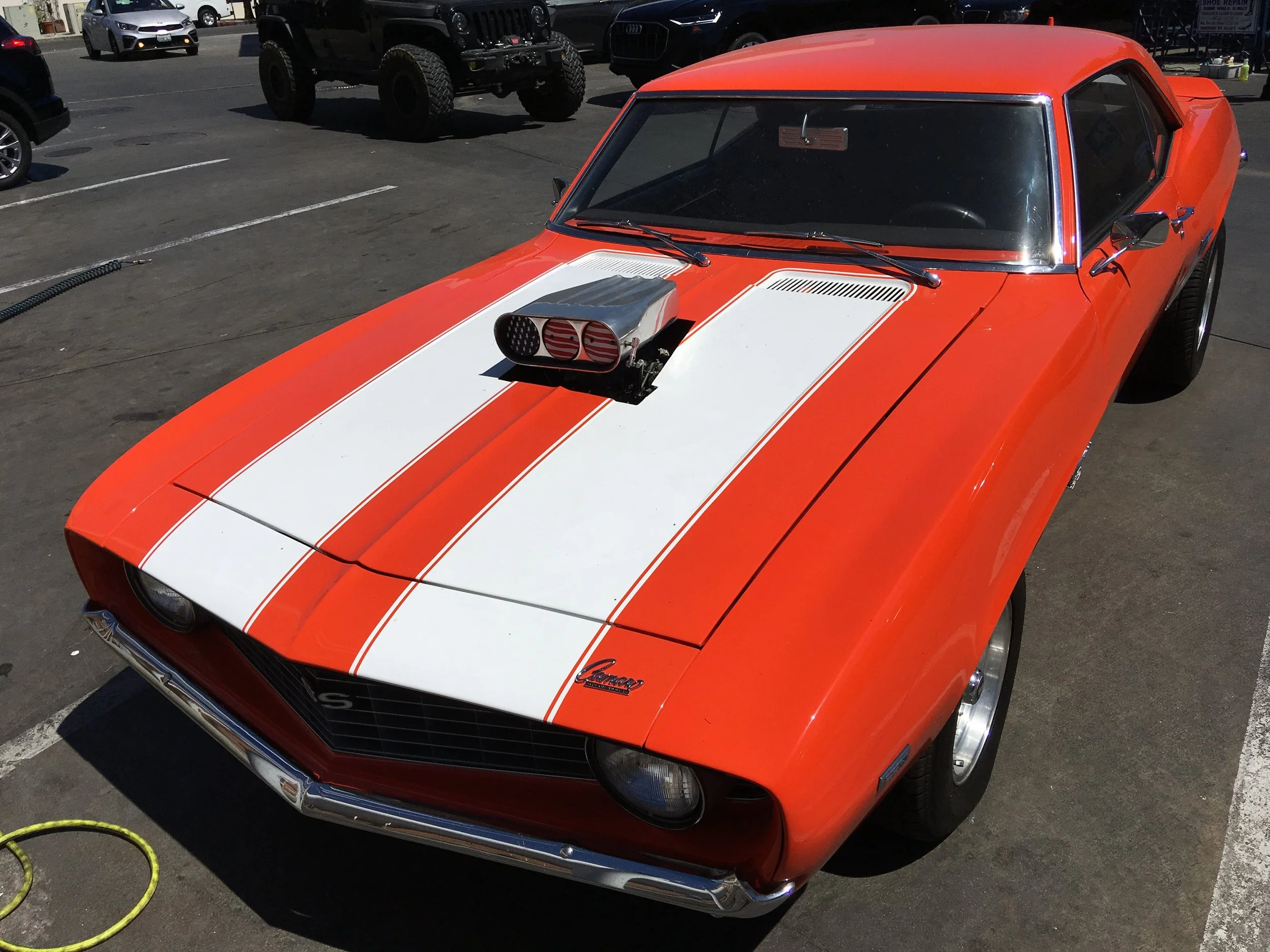 Orange vintage Chevrolet Camaro with white racing stripe, custom blower on the hood, parked in a lot with other vehicles.