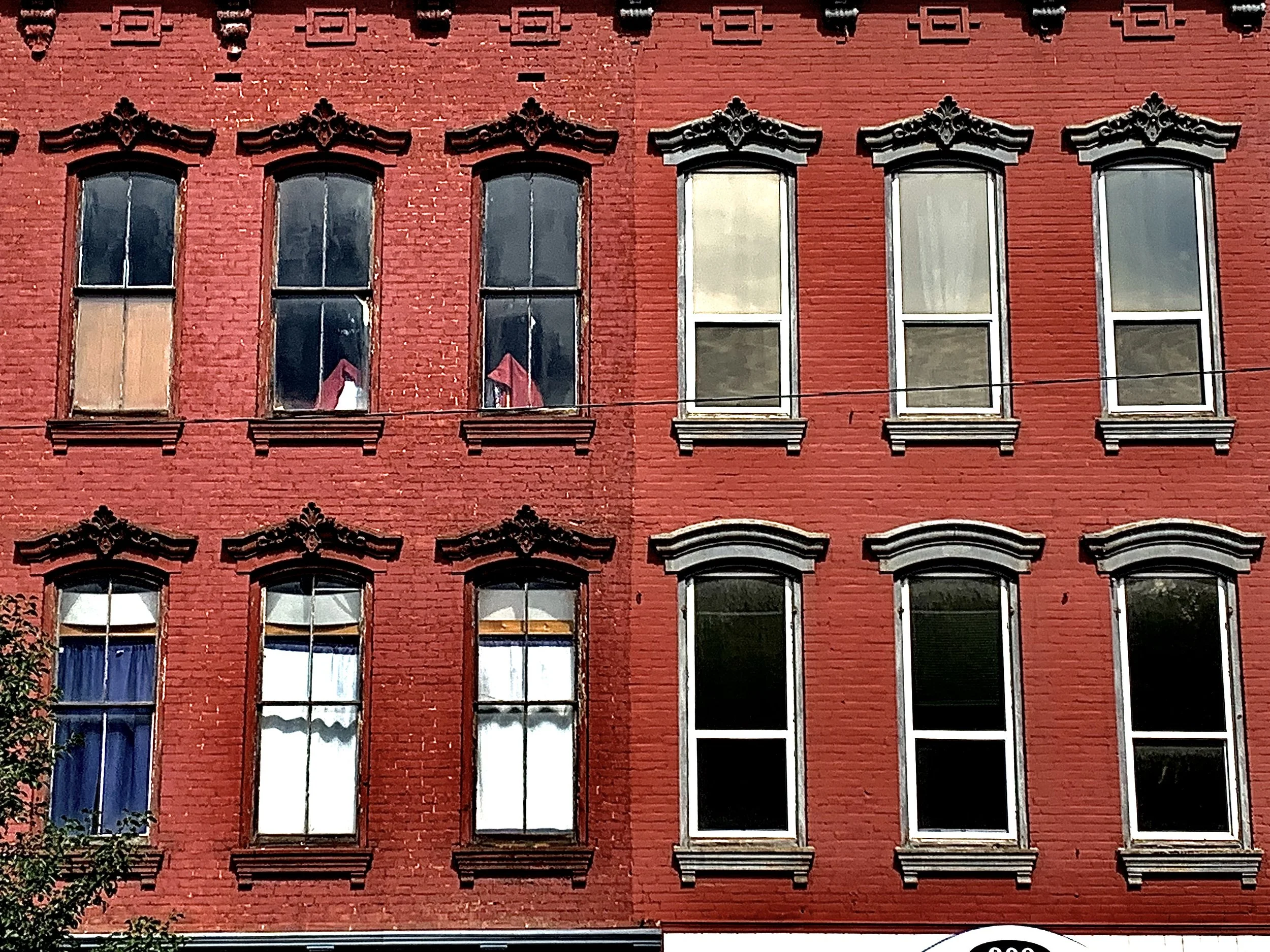 A red brick building with two floors, featuring nine windows with white frames and decorative black trim at the top, some with curtains visible inside.