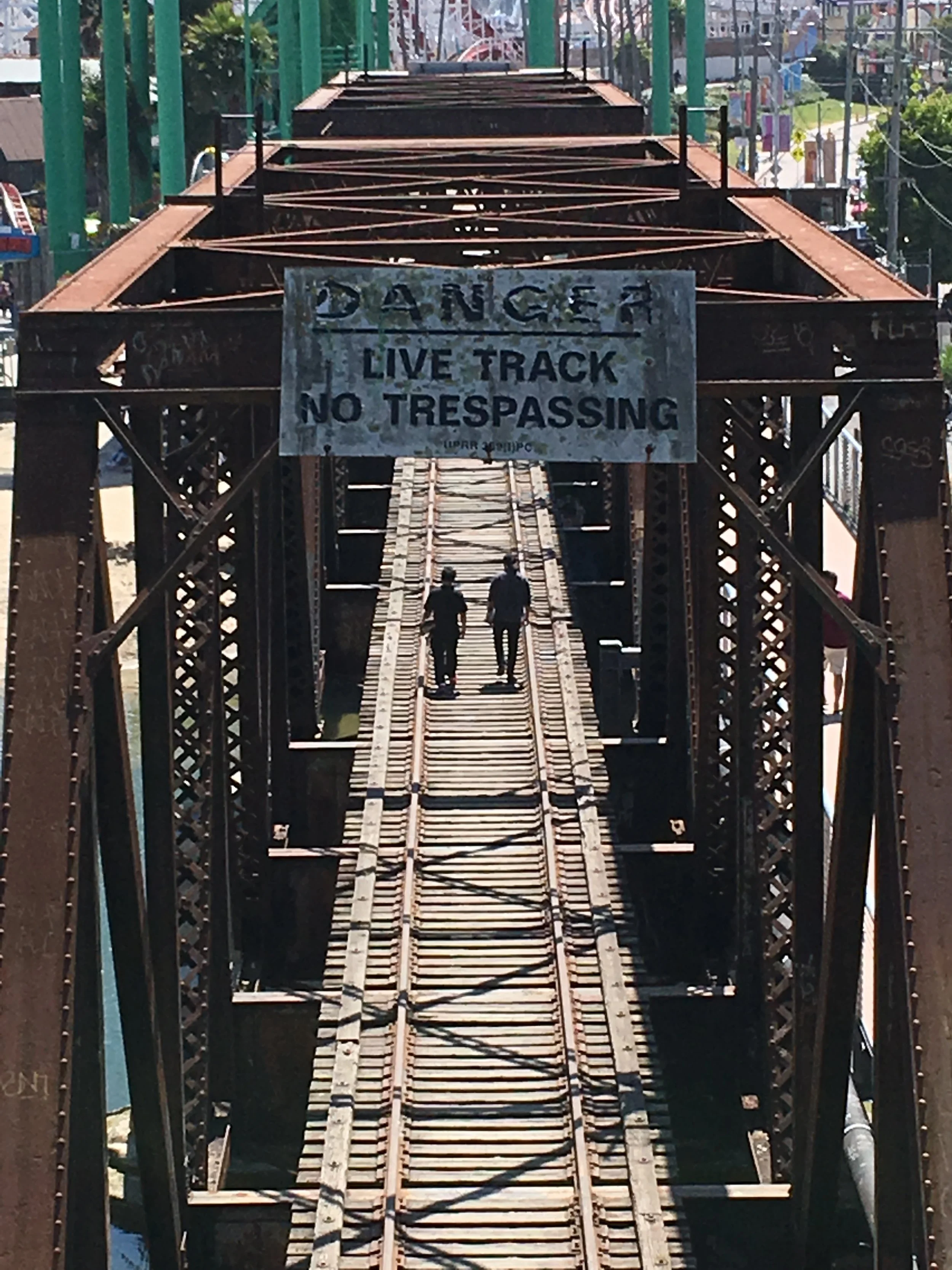 An old, rusty train bridge with a warning sign that says 'DANGER LIVE TRACK NO TRESPASSING'. Two people are walking across the wooden planks of the bridge.
