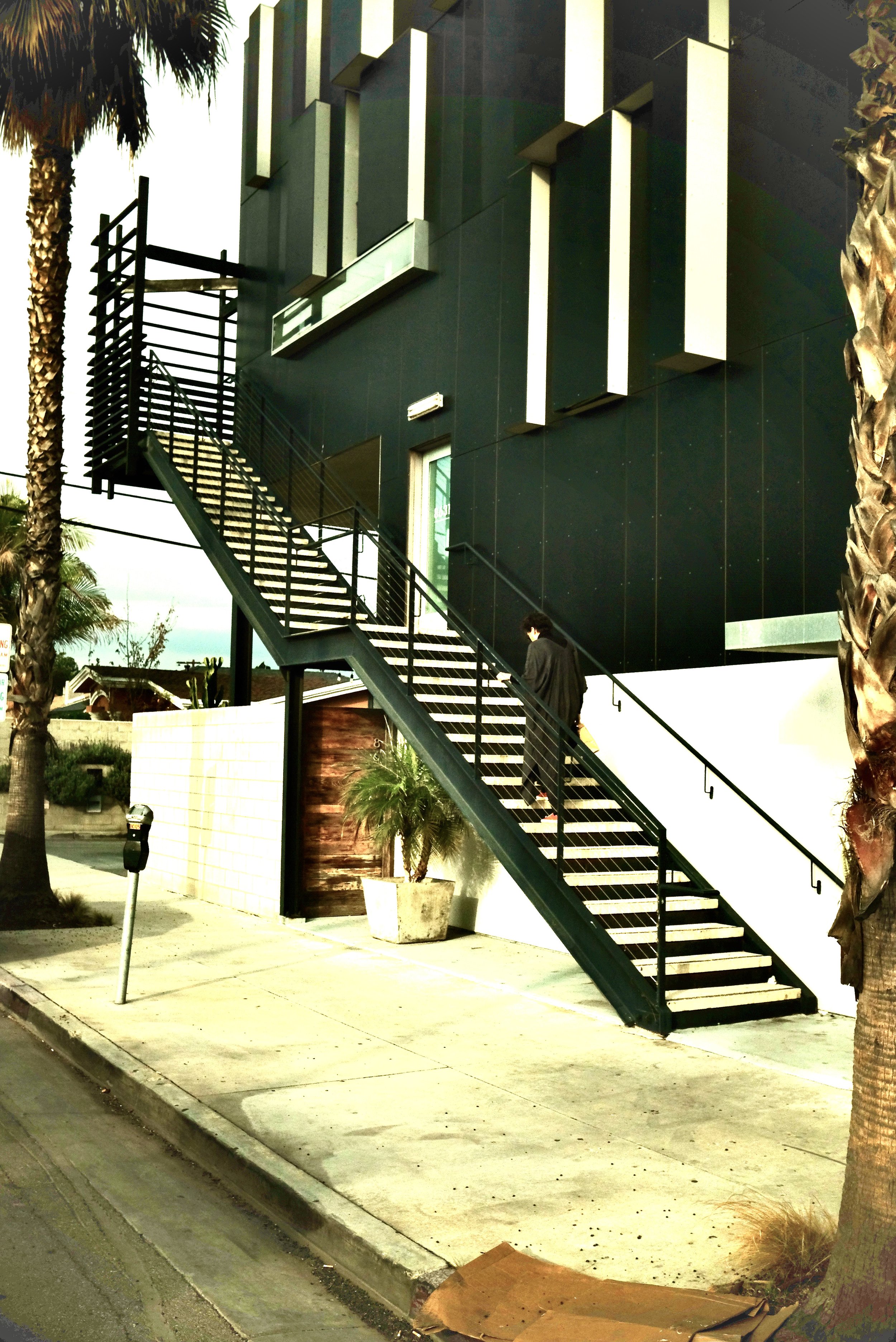 Modern black building with large vertical rectangular accents, exterior staircase with black railings, palm trees, concrete sidewalk, and a person walking down the stairs.