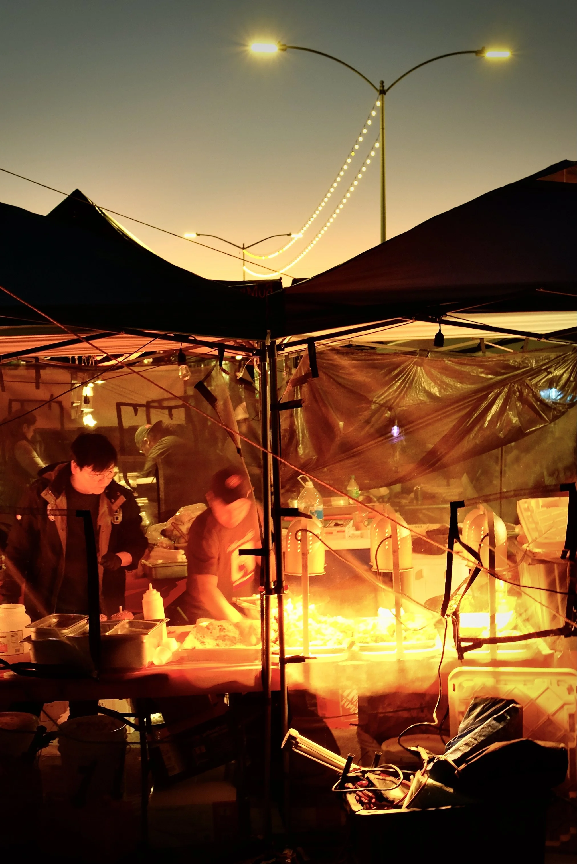 A busy outdoor food stall at sunset with people preparing food under a canopy, illuminated by warm lights and a bright cooking fire.