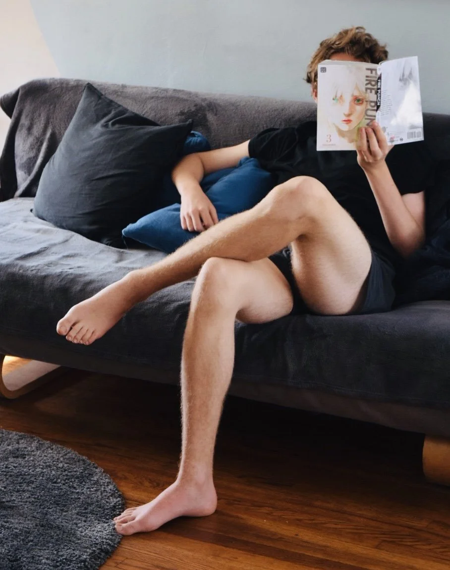 Person lying on a couch with legs crossed, reading a magazine, with pillows behind them and hardwood floor beneath.