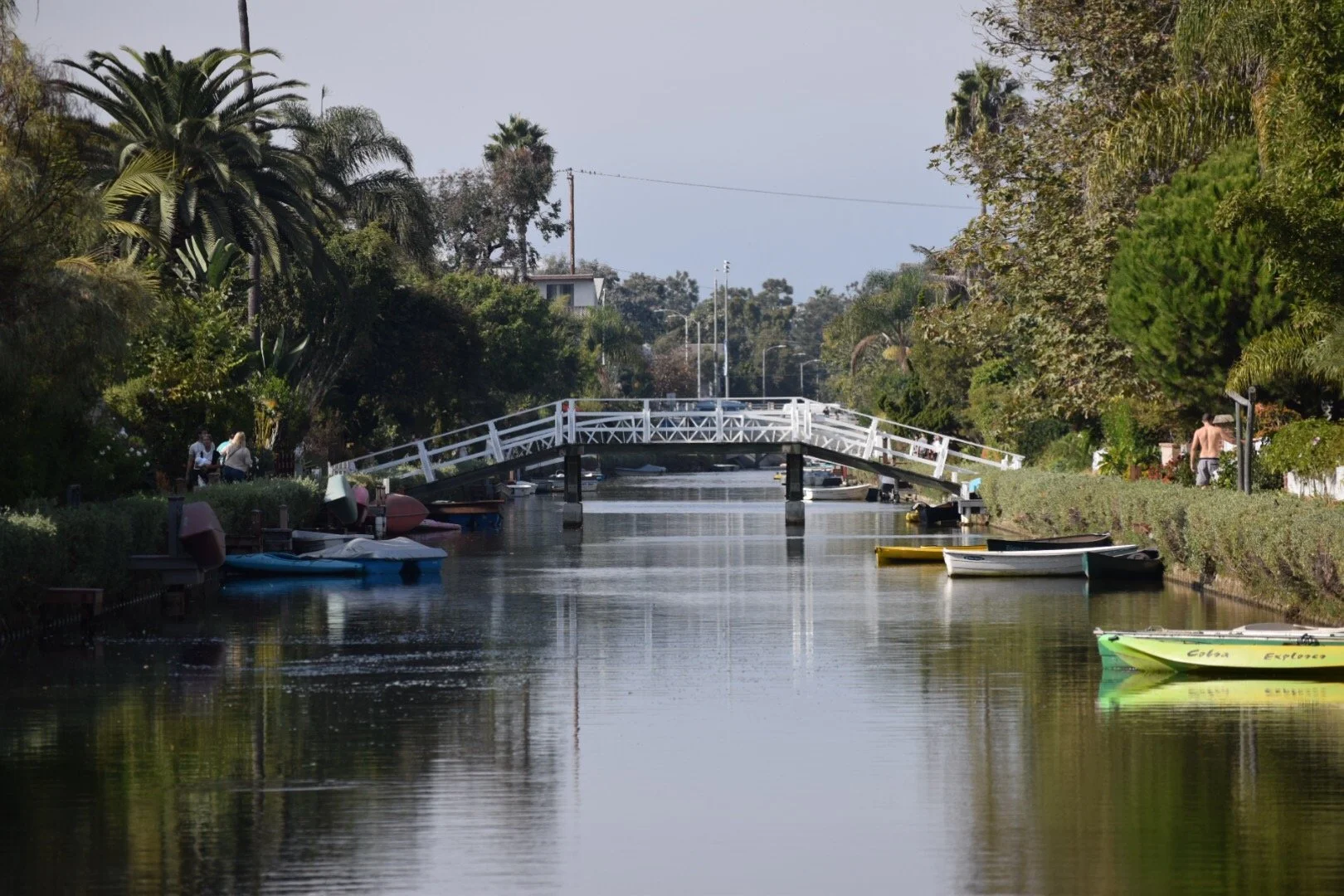 A canal with boats on the sides, a white arched footbridge in the middle, and green trees lining both banks.
