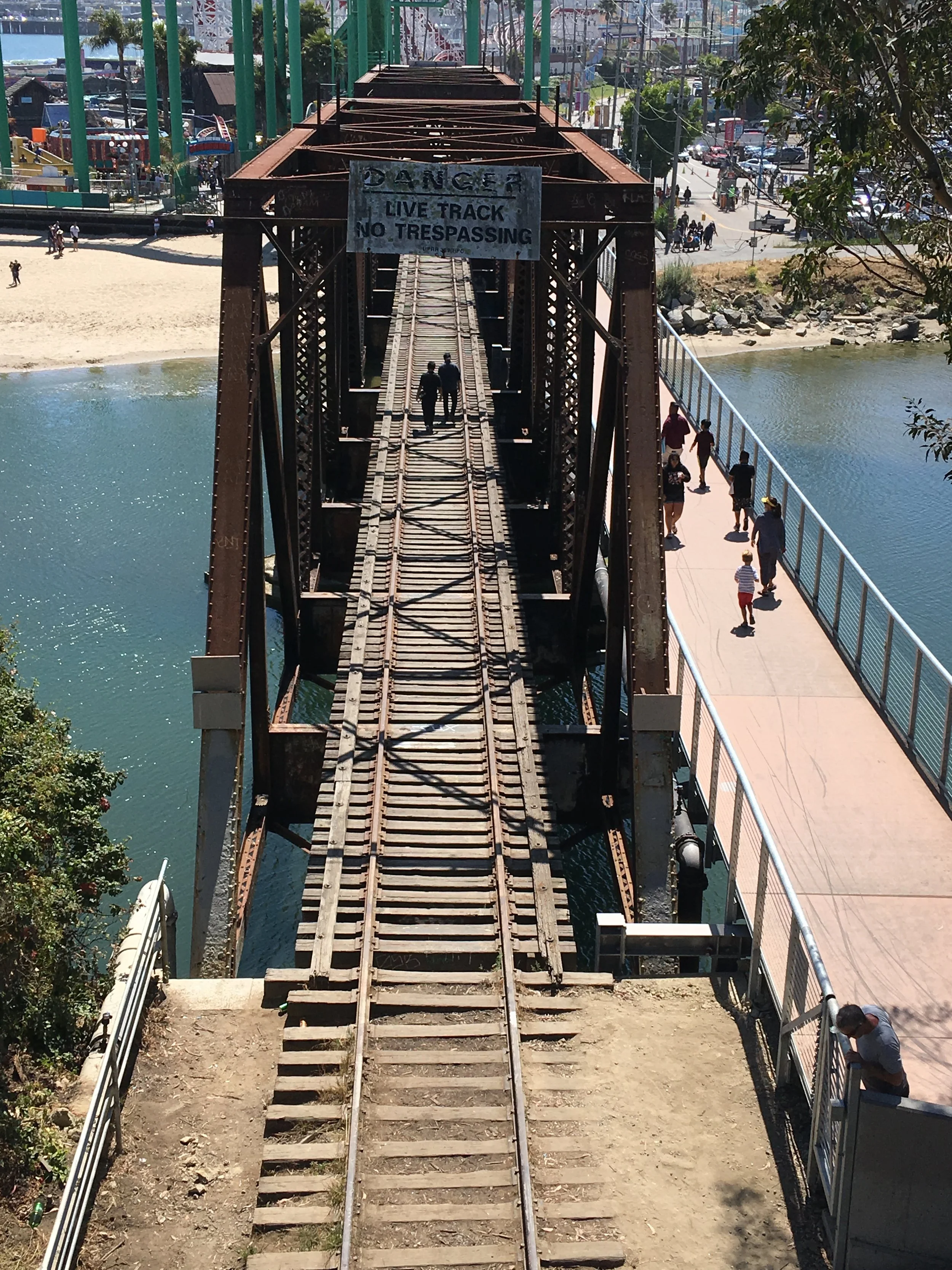 An old, rusty train track bridge over water with a warning sign that says "Danger: Live Track No Trespassing." Pedestrians walk along a pathway beside the water on a bright, sunny day.