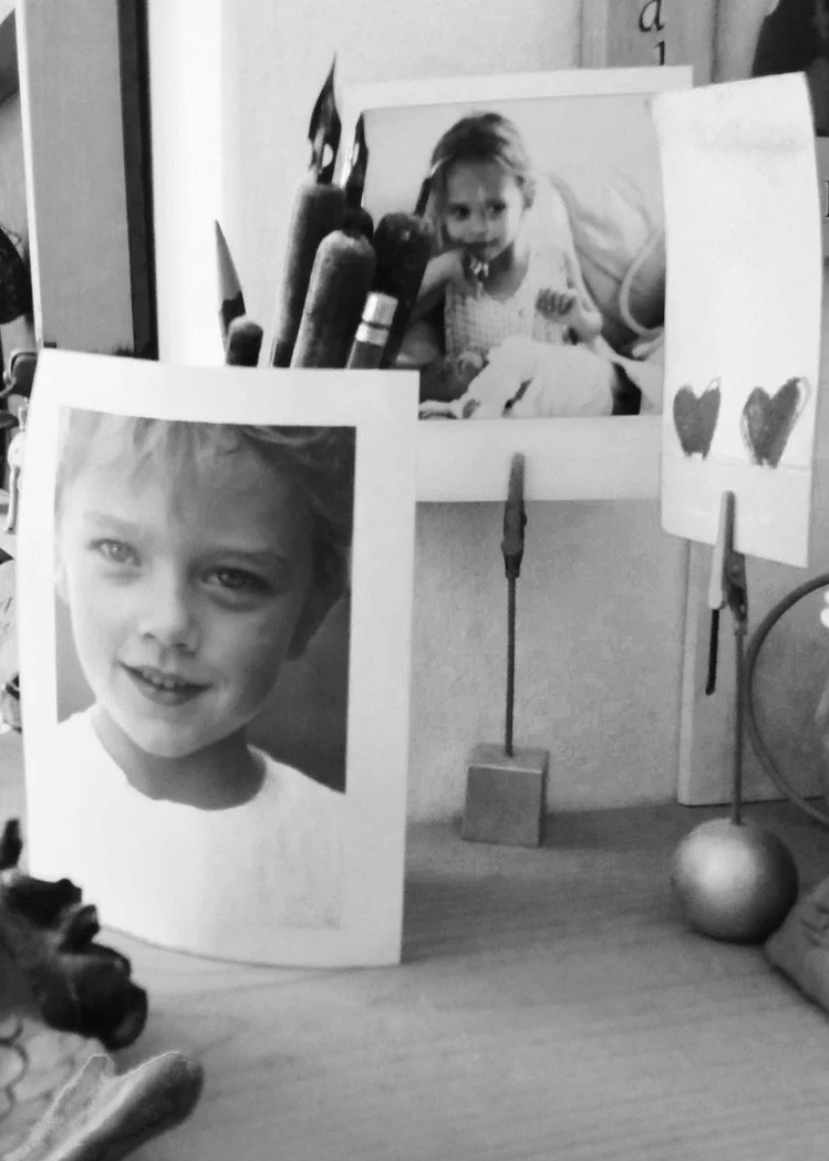Black and white photo display featuring a large portrait of a young boy with light-colored hair, a picture of a girl lying in a hospital bed, and a drawing of two hearts, all on a desk with pencils, a stress ball, and a pen.