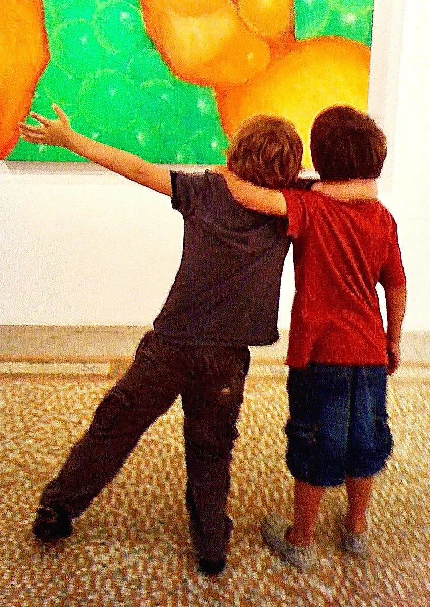 Two young boys are standing together with their arms around each other, looking at a colorful painting in an art gallery. One boy extends his arm toward the painting.