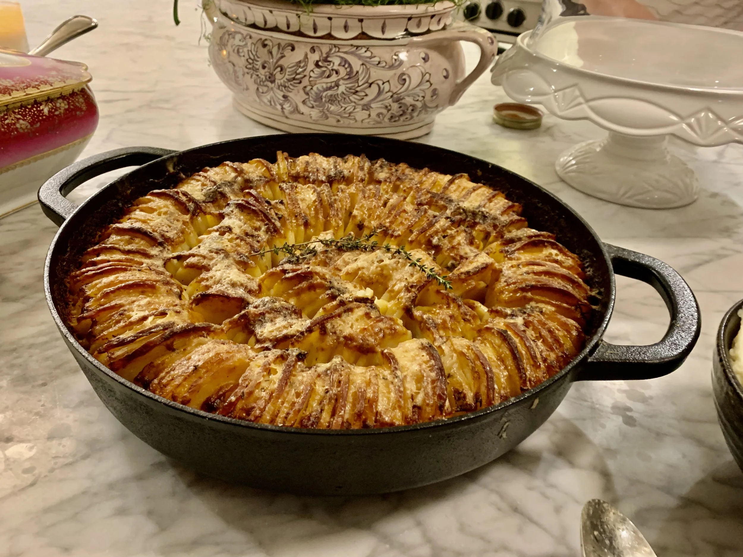 A baked dish of sliced potatoes arranged in a circular pattern in a black cast iron skillet, garnished with a sprig of thyme and topped with grated cheese.