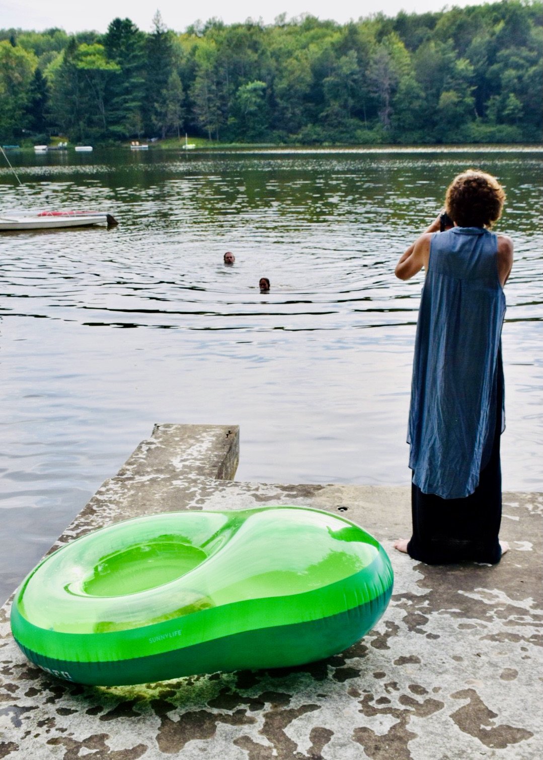 A person standing on a riverbank taking a photo of two people swimming in the water, with a green inner tube on the ground nearby, and boats and lush trees in the background.