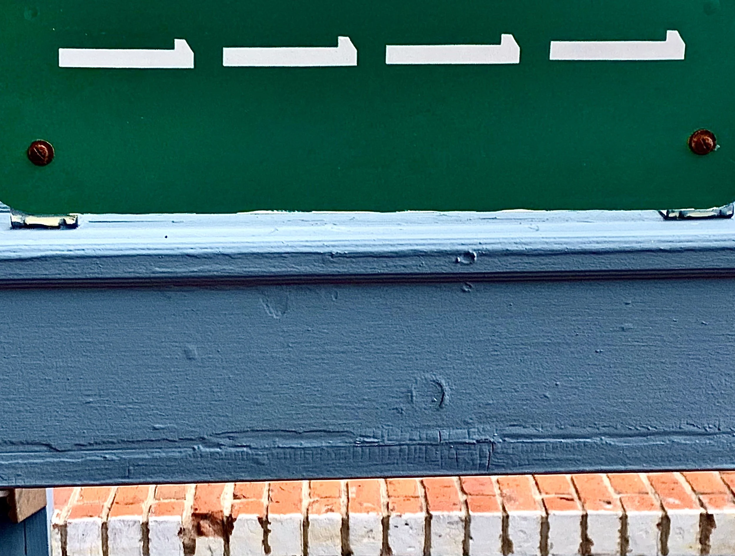 Close-up of a green mailbox with white letter slots mounted above a blue wooden ledge, sitting in front of a brick wall.