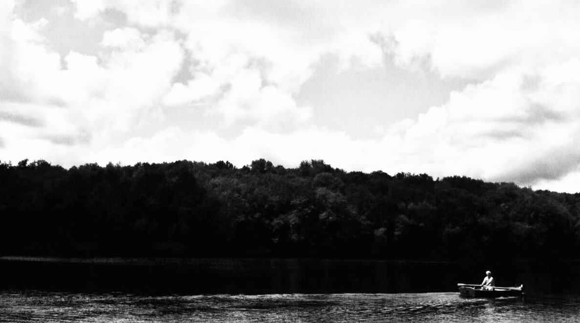 A person rowing a boat on a calm river with a dense forested hill in the background and cloudy sky above.