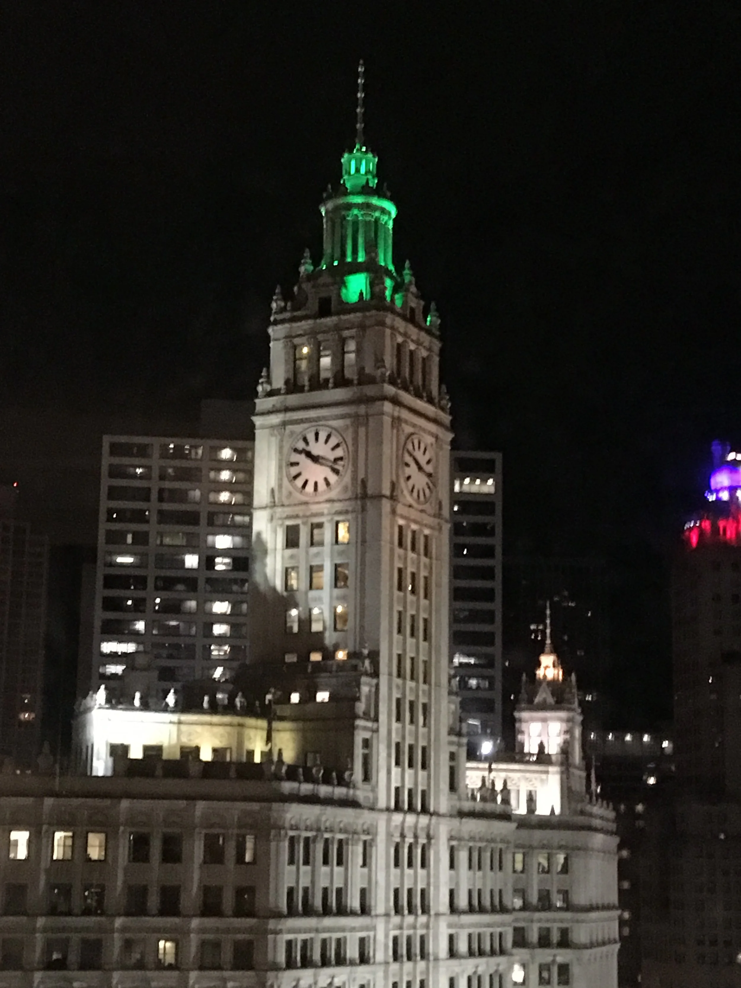 Nighttime cityscape with a tall clock tower illuminated with green lights at the top, surrounded by modern buildings.