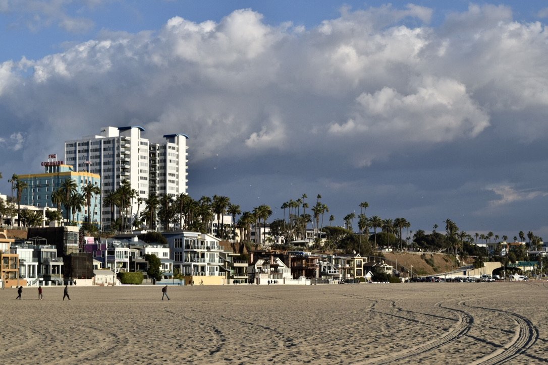 People walking on a sandy beach with modern buildings and palm trees in the background under a partly cloudy sky.