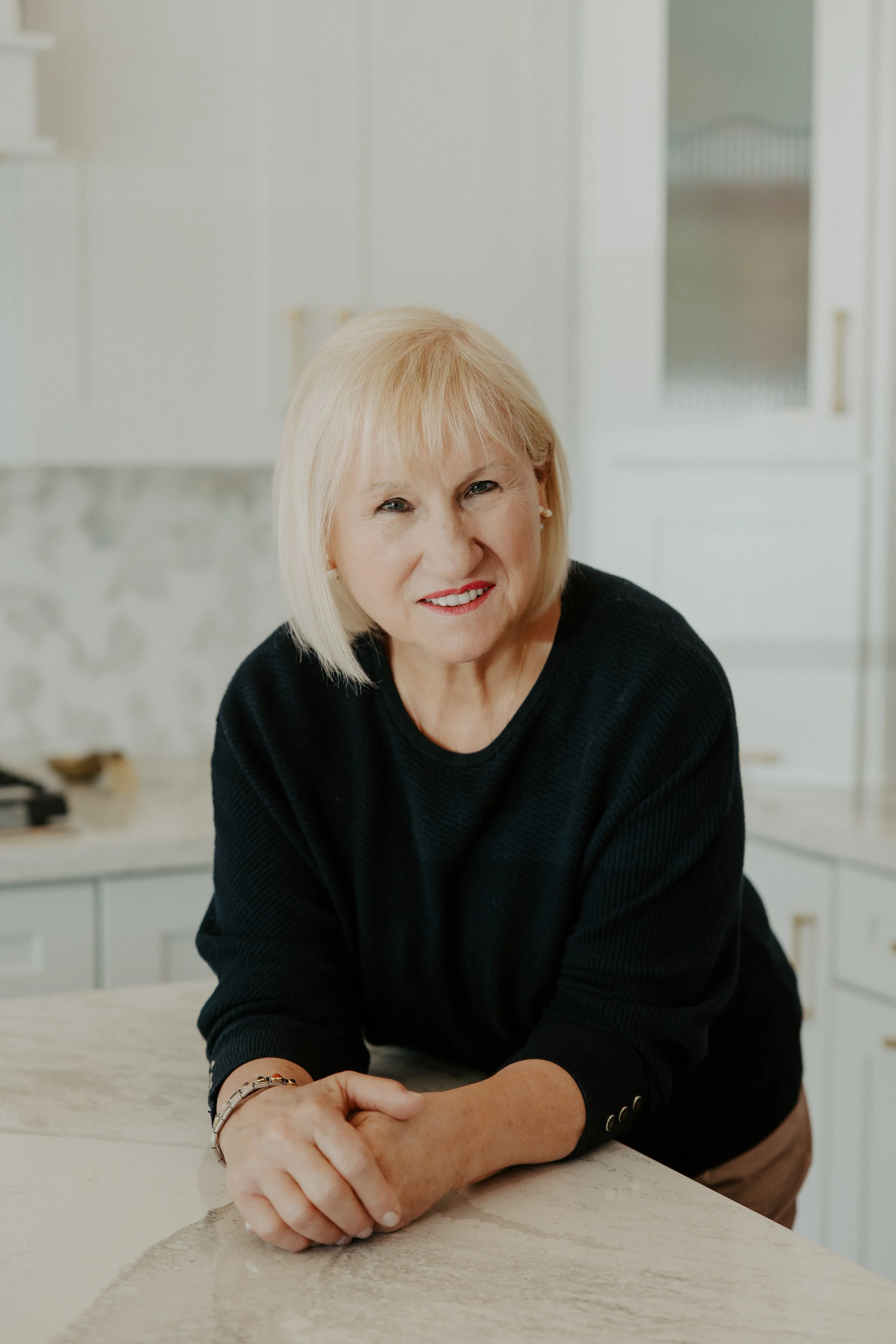 a smiling woman standing in a white kitchen leaning on a taj mahal countertop