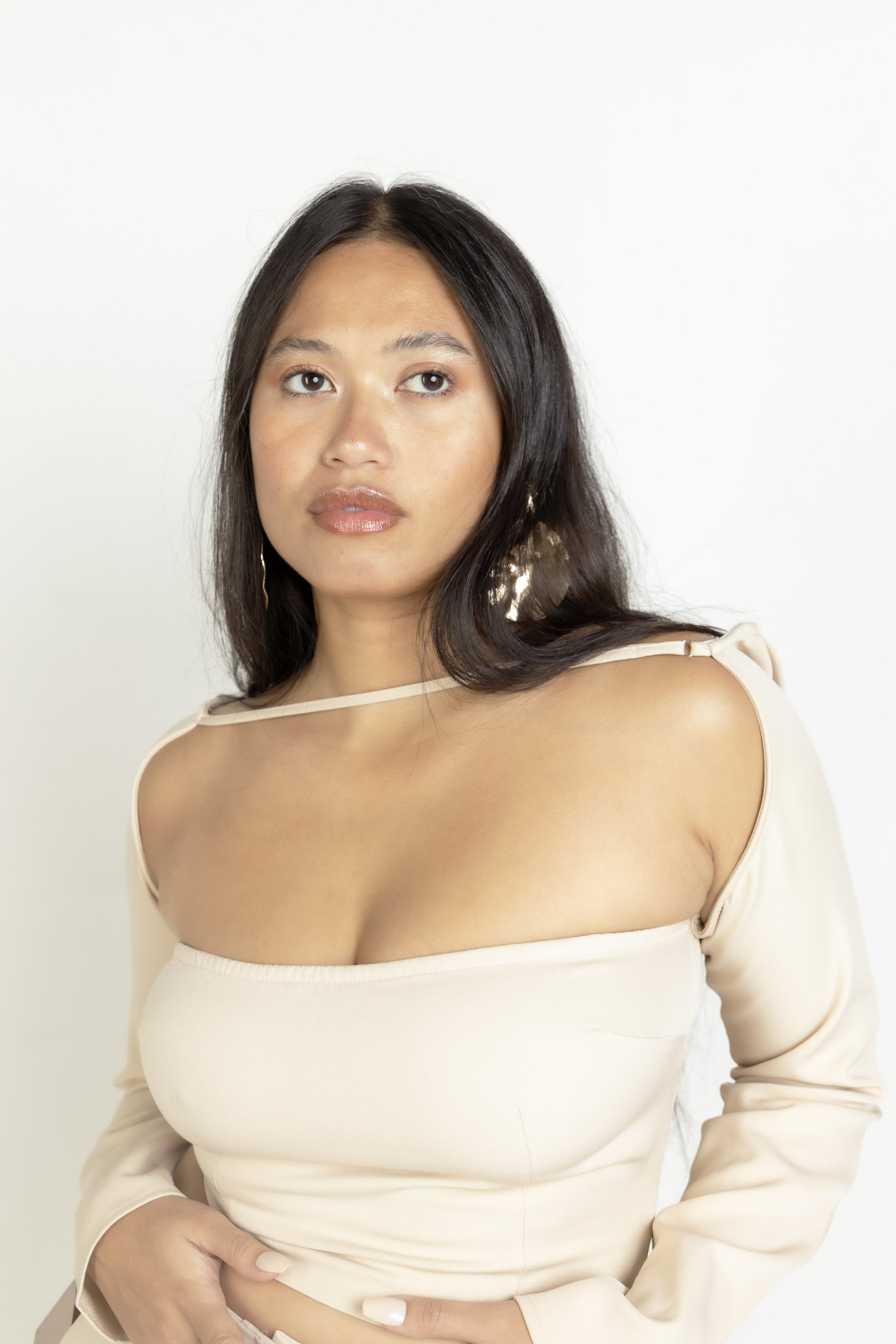 A woman with shoulder-length dark hair wearing a beige off-the-shoulder top with cutout details, posing against a white background.