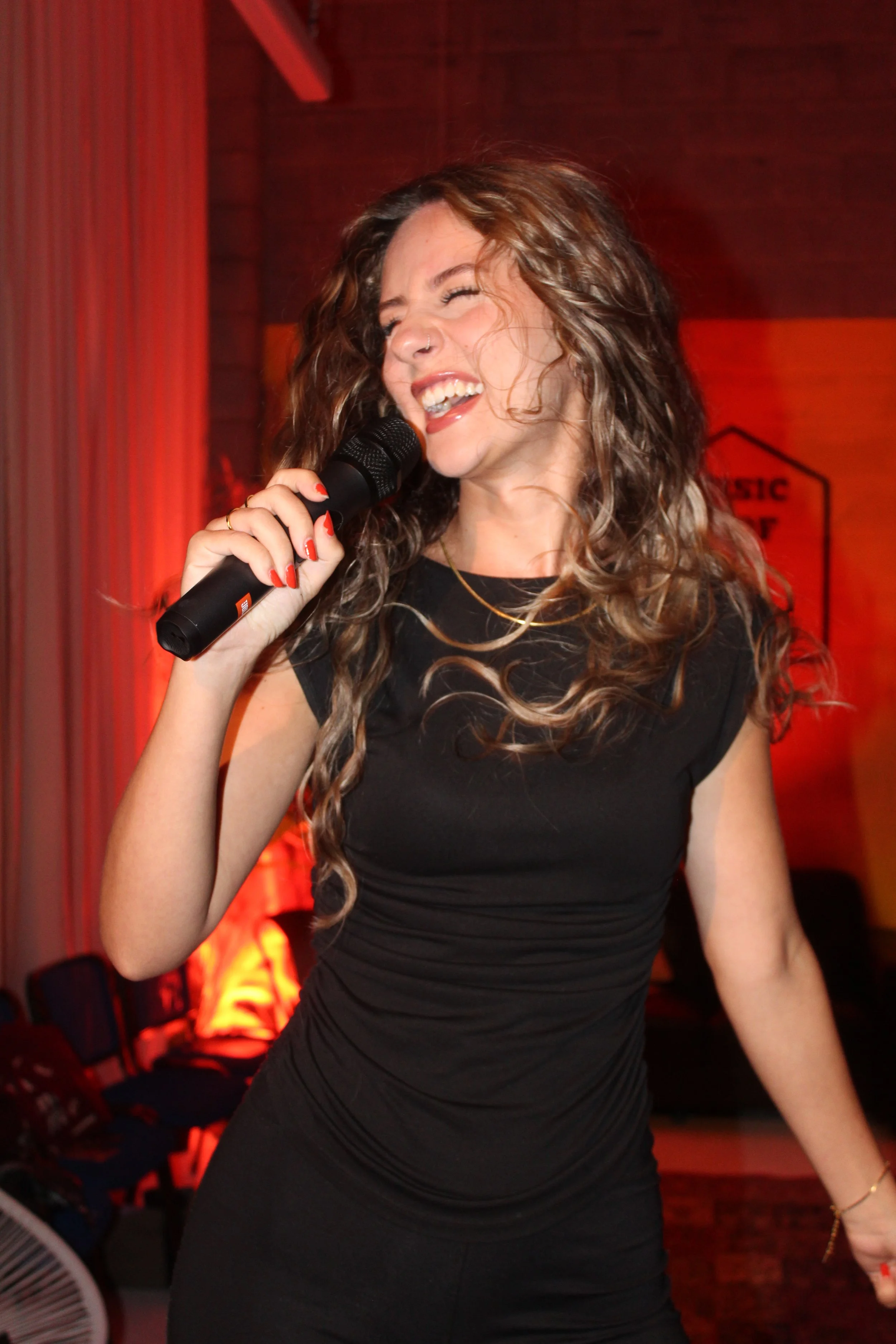 A woman singing and holding a microphone, with curly hair, wearing a black dress, in a room with warm lighting.