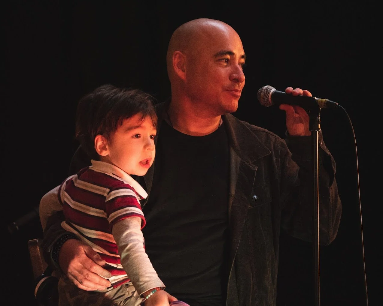 A man holding a young child while speaking into a microphone on a dark stage.