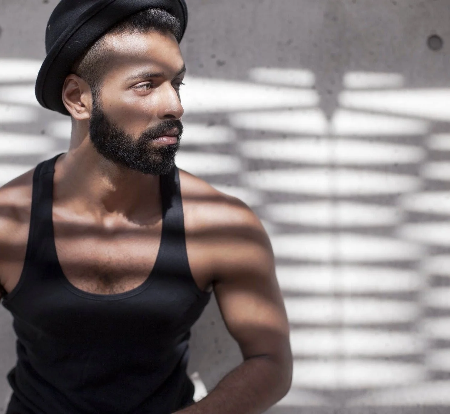 A man with a beard and short hair wearing a black tank top and a black hat looks to the side. Shadows of blinds are cast on his face and shoulder against a gray wall.