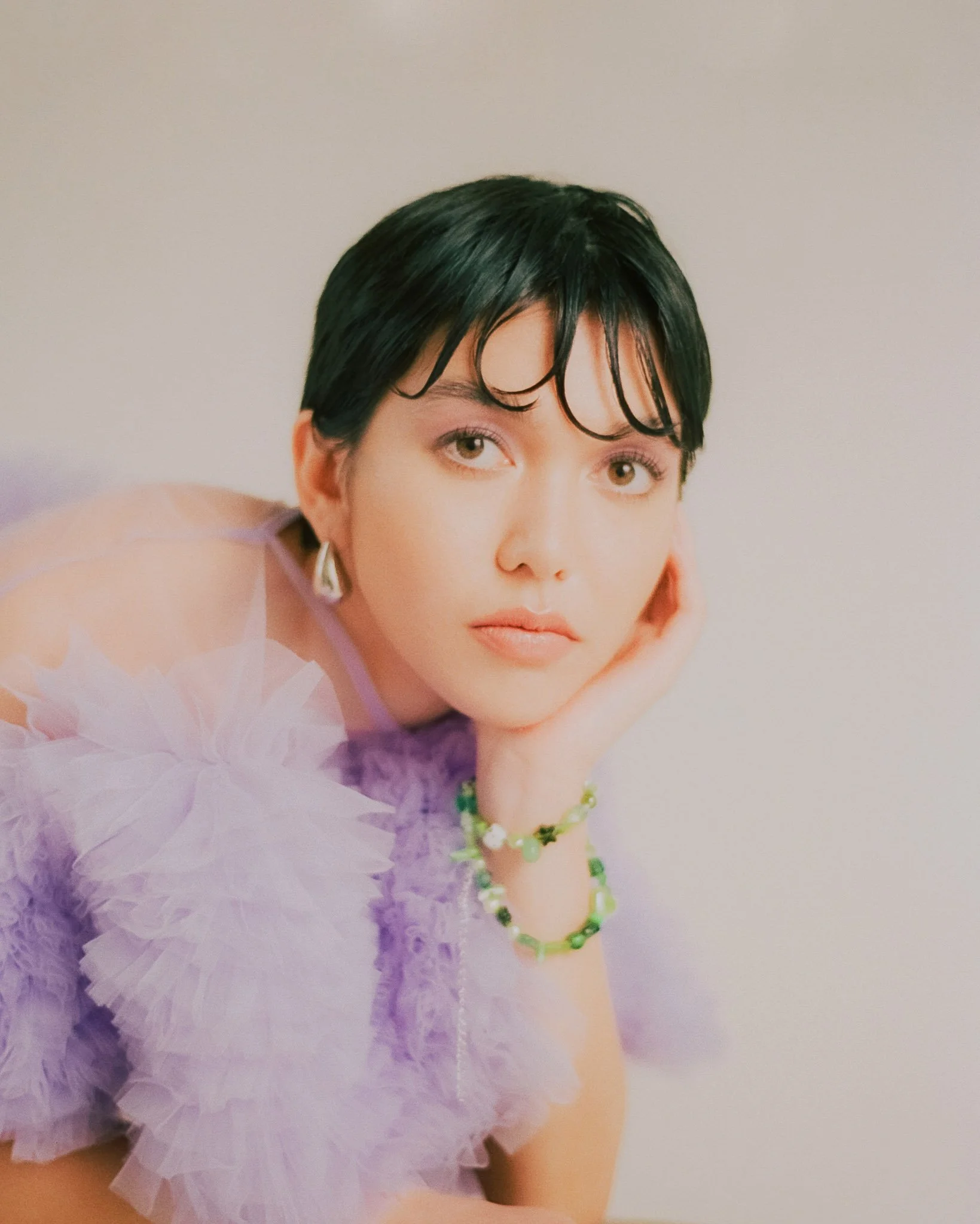 A woman with short black hair, wearing a light purple ruffled dress and green jewelry, resting her chin on her hand, looking at the camera with a neutral expression.