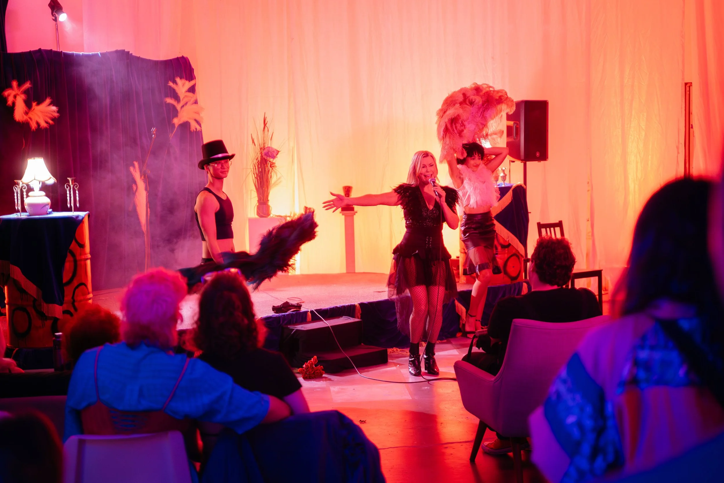 A lively stage performance with performers in costumes, one singer with a microphone and another with a large feathered headdress, in front of an audience in a warmly lit venue.