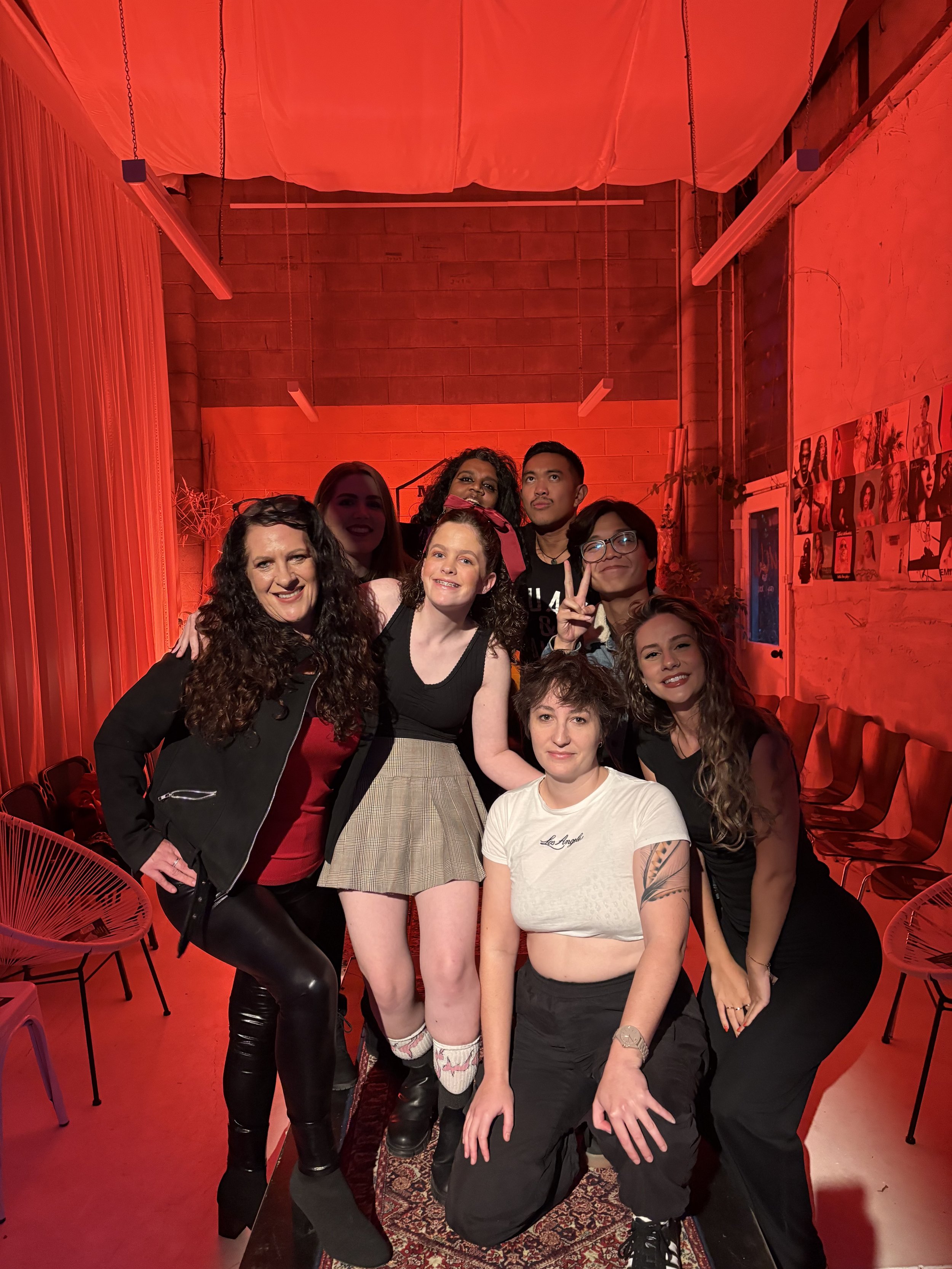 Group of seven women and one man posing together in a room with red lighting, decorated with photos on the wall and chairs along the sides.